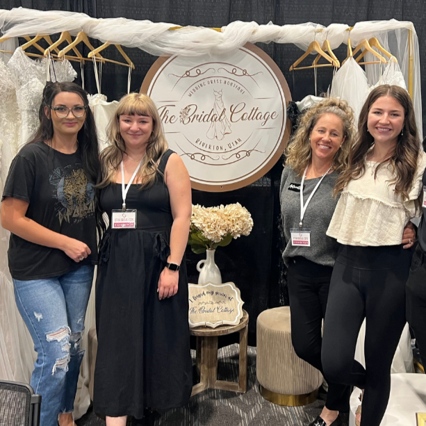 Four women standing together inside a bridal shop named The Bridal Cottage, with bridal dresses hanging behind them and a round sign above that reads The Bridal Cottage, RiverJorn, Utah. There is a vase of flowers on the small table in front of them.