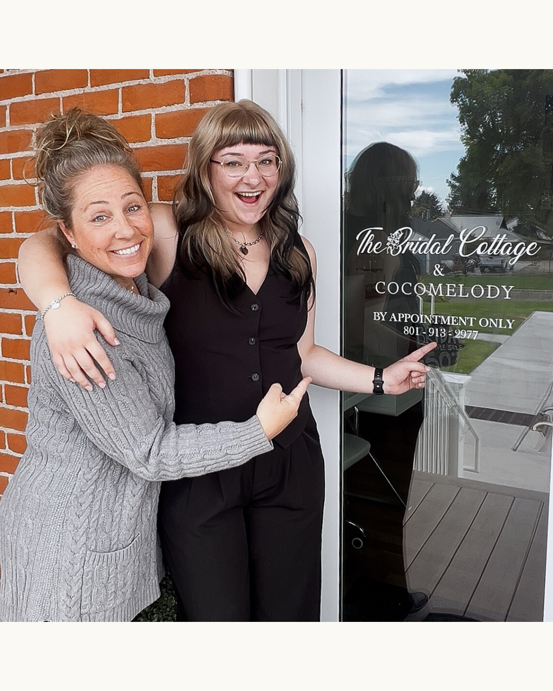 Two women smiling and pointing at a glass door sign for The Bridal Cottage and Cocomelody, located in a brick building, with a reflection of outdoors in the glass.