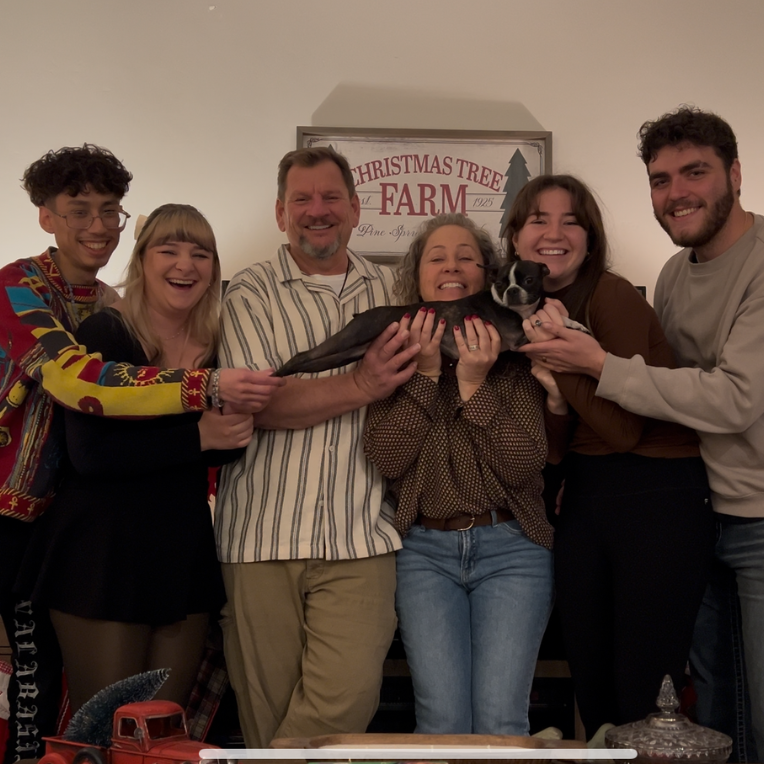 Group of six people holding a small black dog in front of a sign that says "Christmas Tree Farm 1925".