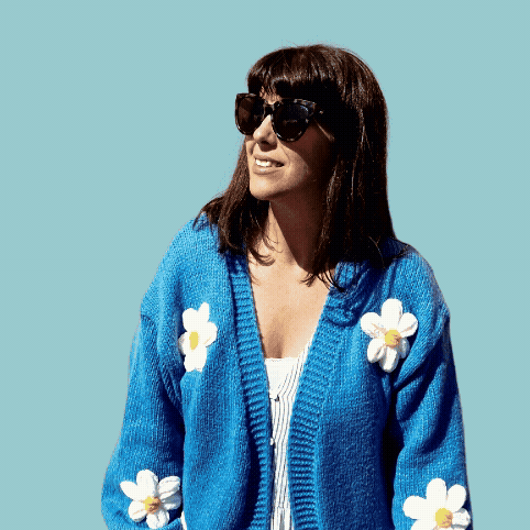 Woman with shoulder-length dark hair wearing sunglasses and a blue cardigan with white flower patterns, smiling against a light blue background.