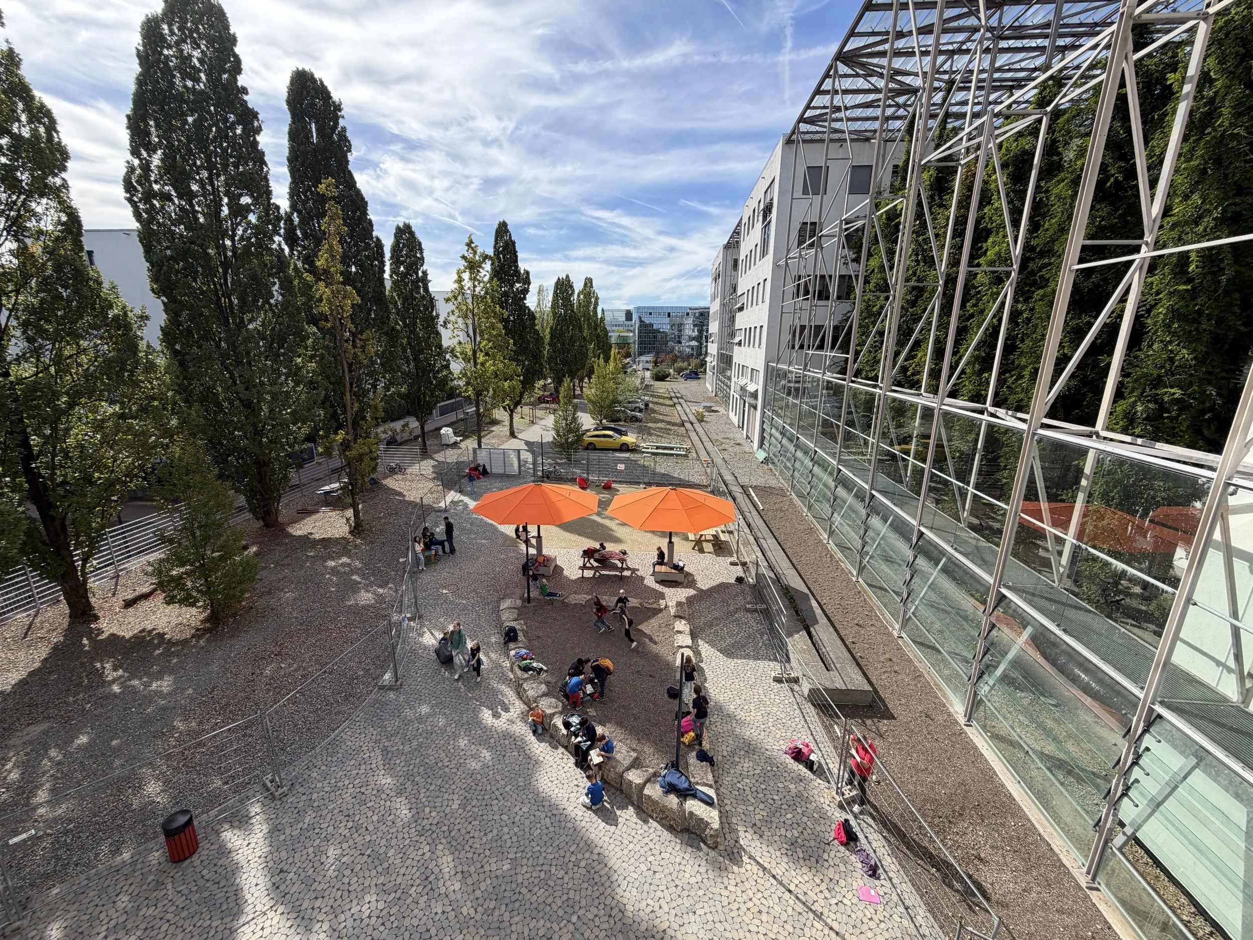 Realisierung im Eiltempo eines Teils des neuen Schulhofs für die ASB Erasmus Frankfurter Stadtschule Grundschule gGmbH bis Schulbeginn 10.8.2026. Bunte Sonnenschirme stehen auf dem von uns oft verwendeten "Arena"-Pflaster. Gelblich davor liegt das Pf
