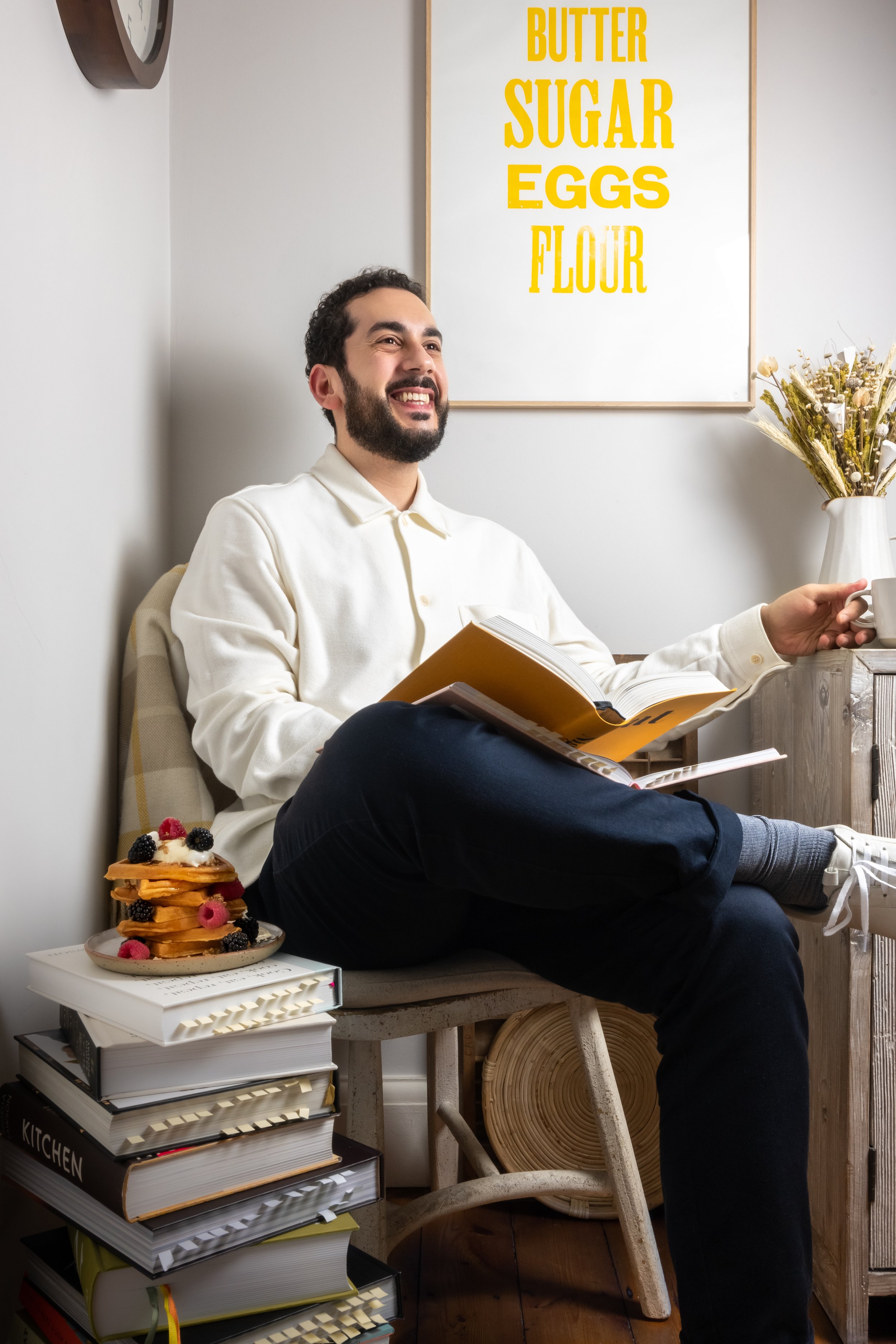 A portrait of photographer, stylist and art director Costas Millas sat in his kitchen studio with a print above him with Butter, Sugar, Flour, Eggs printed in yellow letters. At his side are a large stack of cookbooks and a plate of waffles.