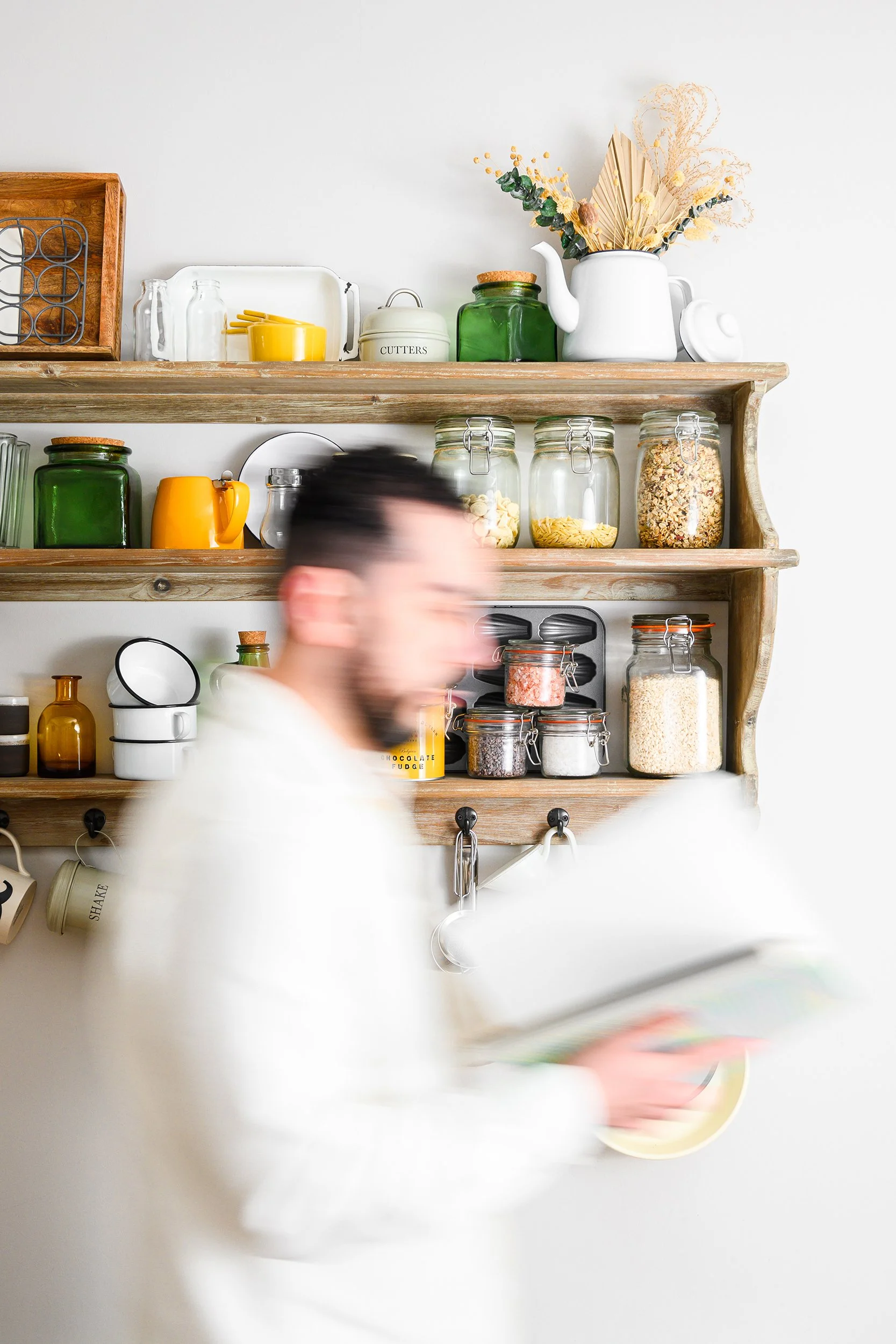 Blurry photo of a man in a white shirt flicking through the pages of a book as he walks past wooden kitchen shelves behind filled with glass jars, cups, and kitchen items which are in focus.