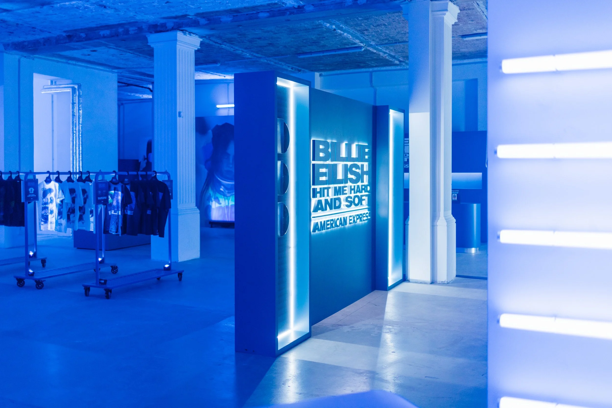 A retail store featuring clothing on racks illuminated with blue lighting, including a lit sign that reads 'BLUE BUSH HIT ME HARD AND SOFT' along with the American Express logo.