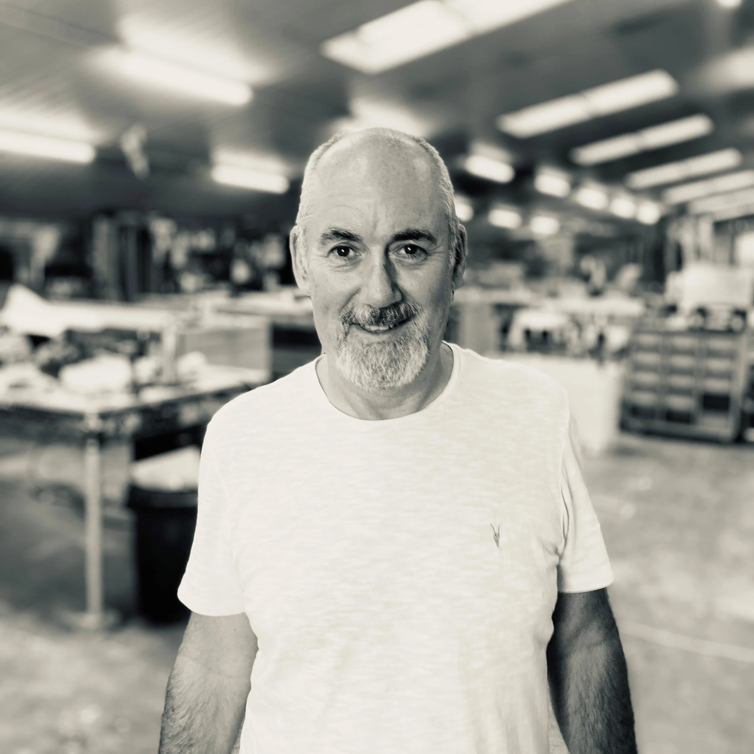 Black and white photo of a man with a goatee, wearing a white shirt, standing in a workshop.