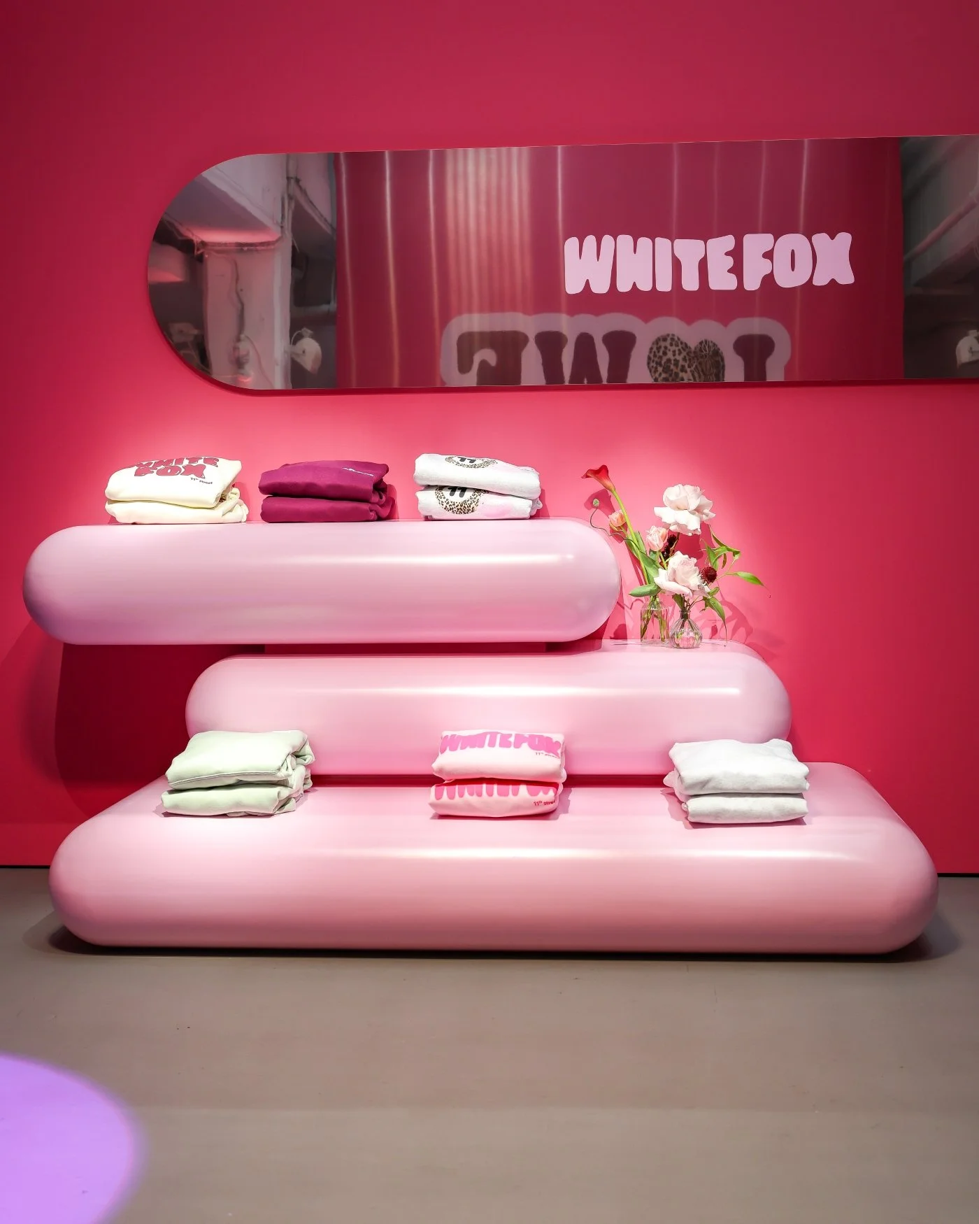 Pink display with folded clothes, flowers, and a mirror reflecting a sign that says 'WHITE FOX' in a retail store.