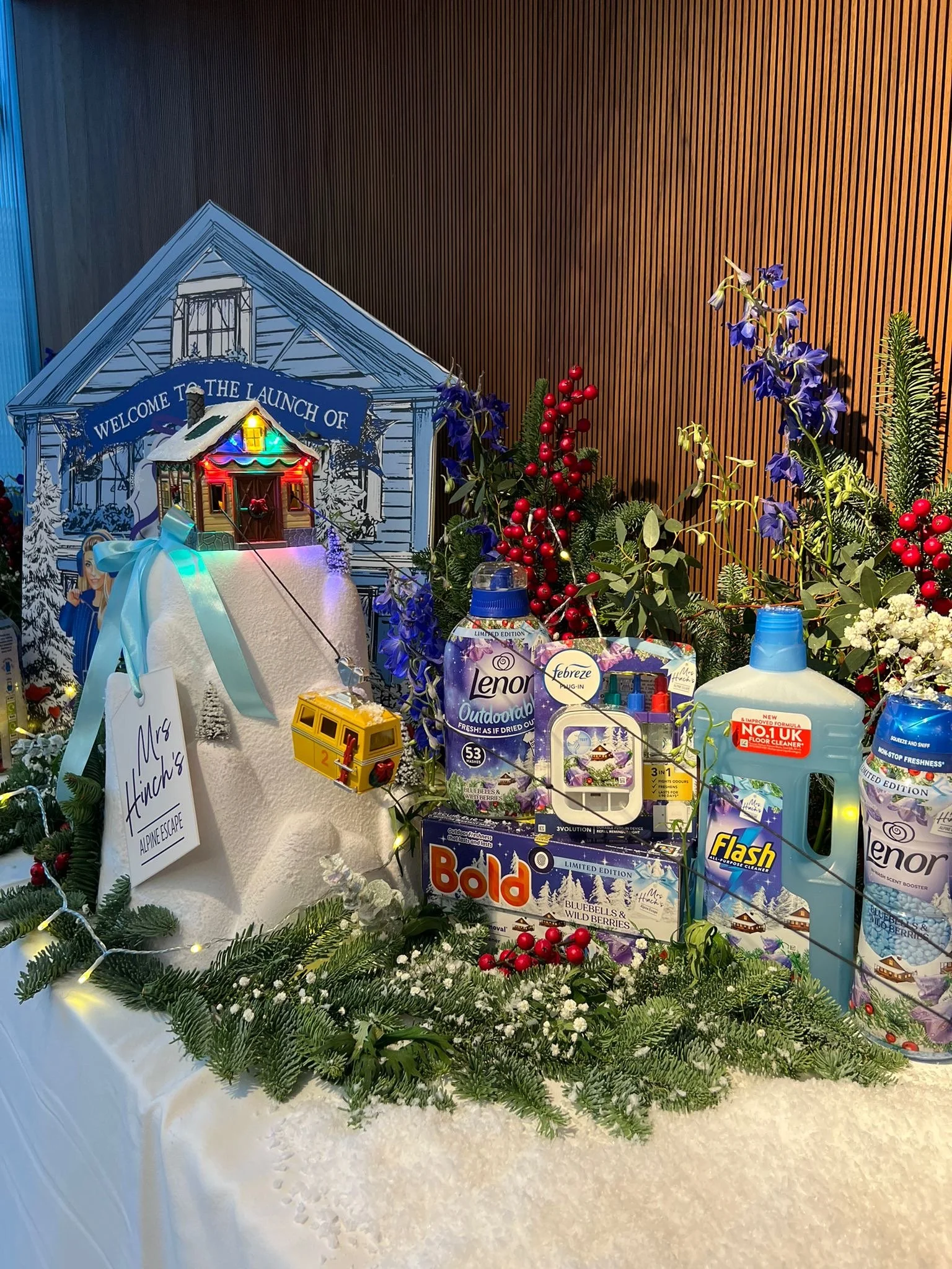 Decorative holiday display featuring a miniature house with colorful string lights, a sign that reads "Welcome to the Launch of" and an illustration of a house, surrounded by greenery, red berries, purple flowers, and various household products such as laundry detergent, floor cleaner, and air freshener, all set against a wooden wall background.