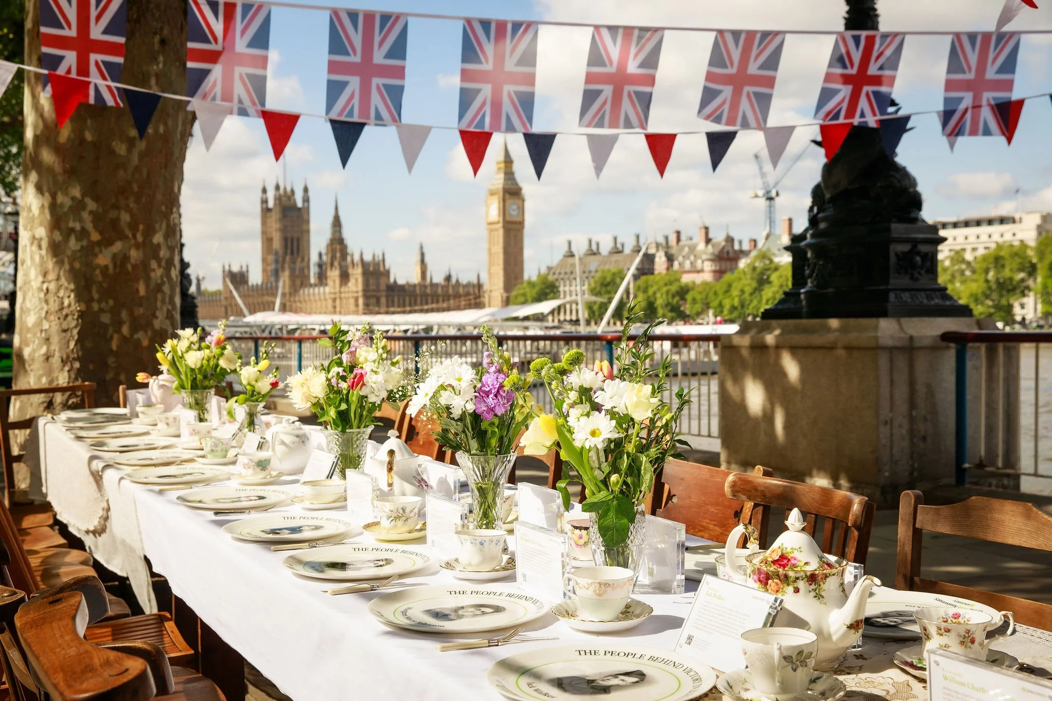A long dining table set with floral centerpieces, teacups, and plates, outdoors in London with Big Ben and the Palace of Westminster in the background, decorated with British flags and bunting.