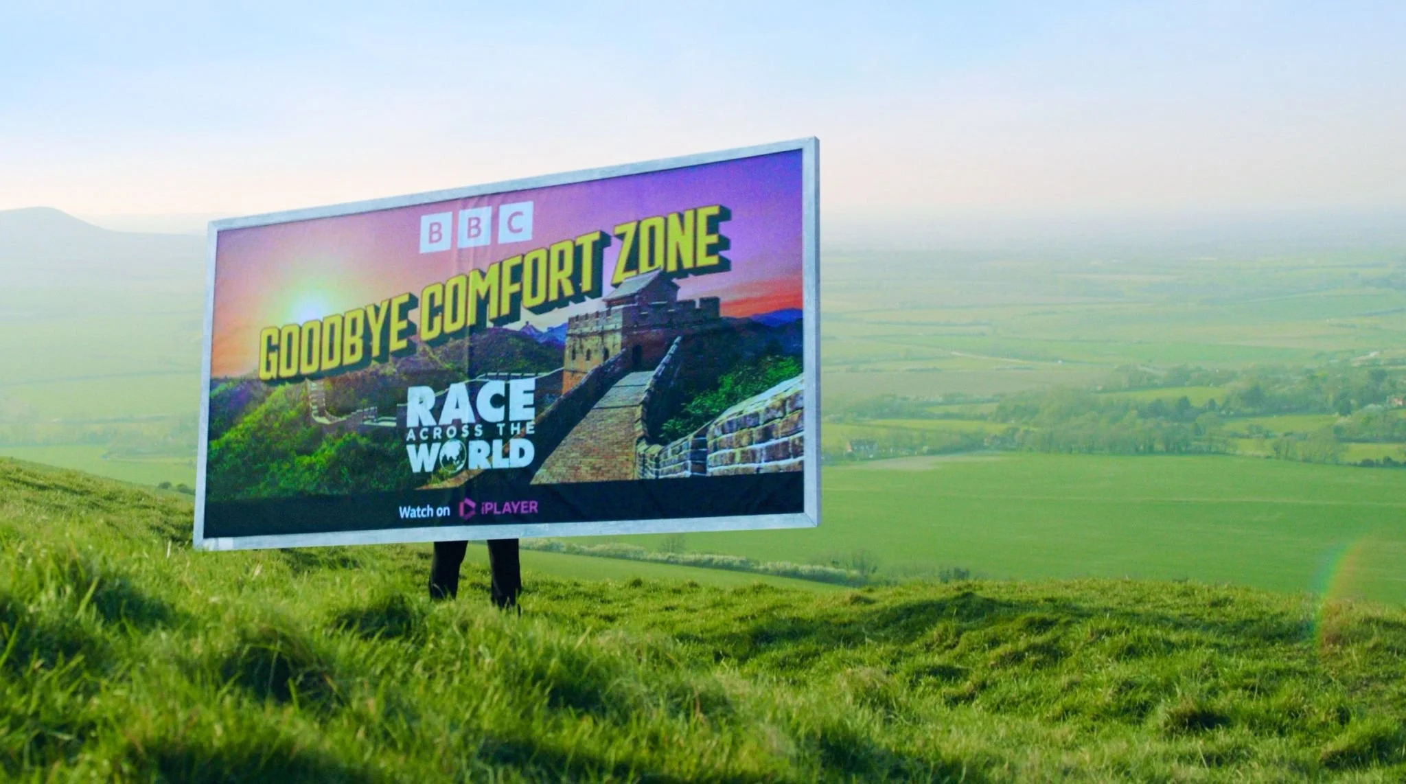 A billboard with the words 'Goodbye Comfort Zone' and 'Race Across the World' in front of a green field and a distant mountain range.