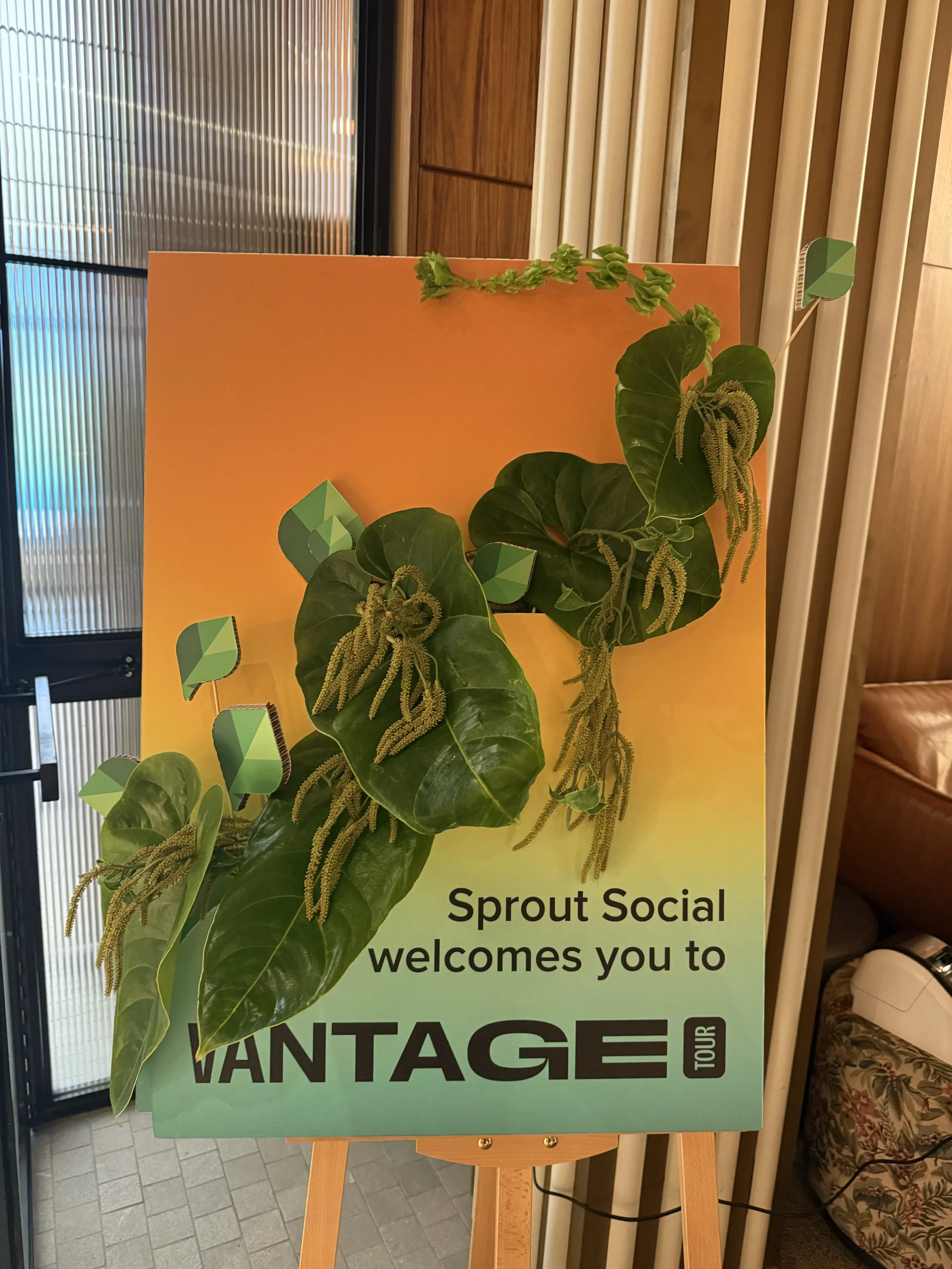 A sign on an easel with a green and yellow gradient background, a large green sprout graphic, and black text that reads, 'Sprout Social welcomes you to VINTAGE Tour.' There are large green leaves and stems attached to the sign, and the background shows part of a wooden wall, vertical blinds, and other interior elements.