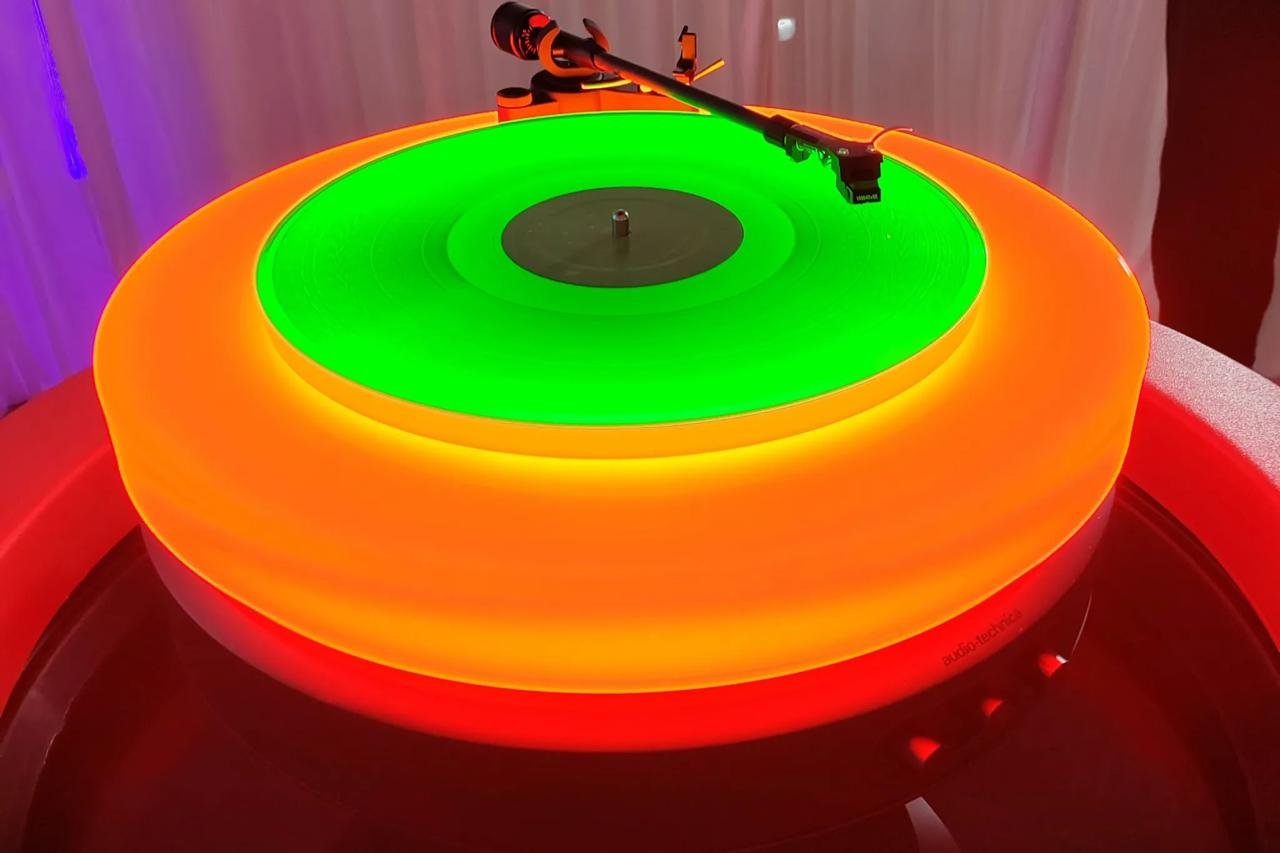 A brightly colored turntable with a neon green and orange glowing platter and a black tonearm.