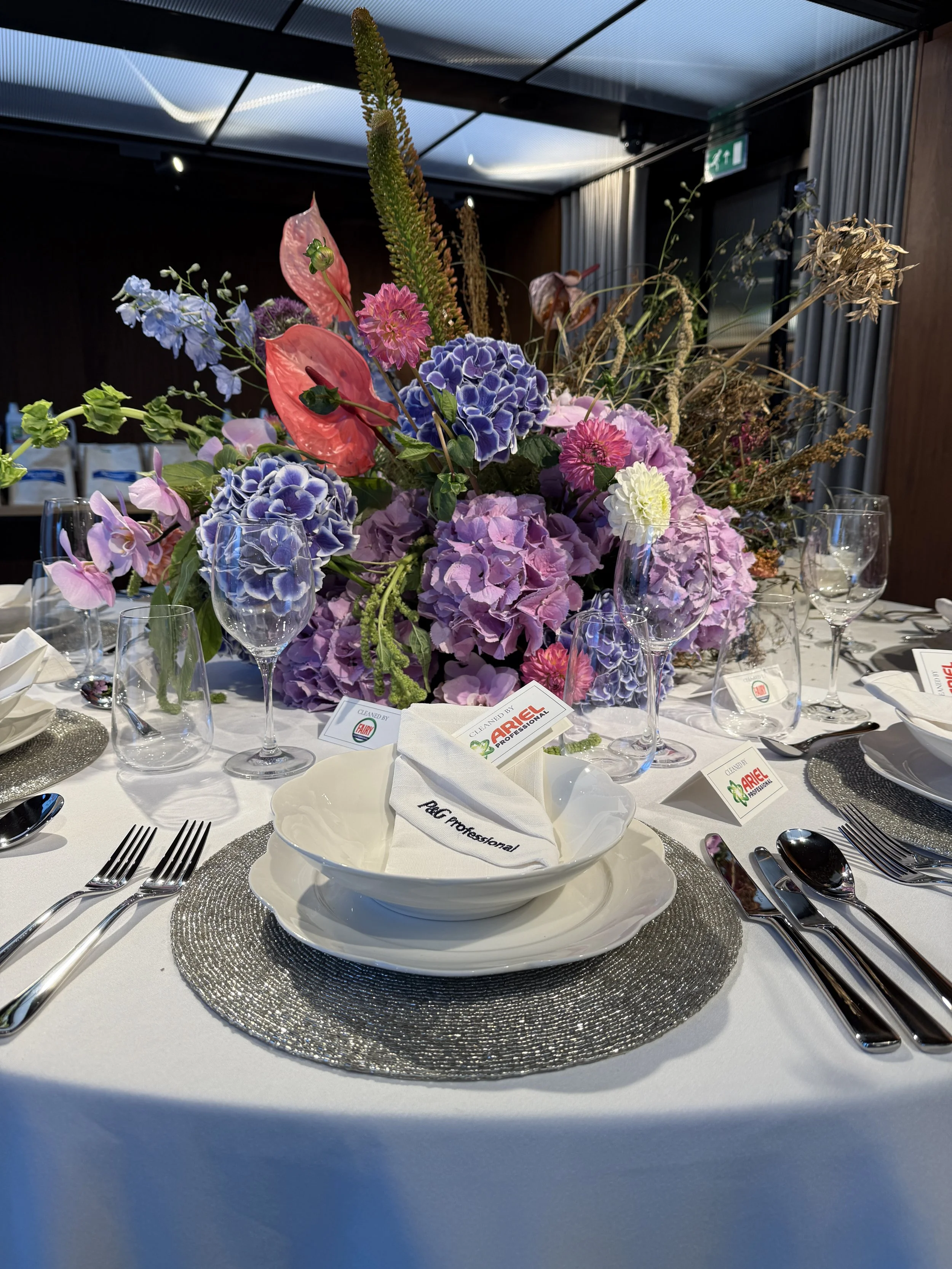 Elegant dining table setup with a large floral centerpiece, white plates, silverware, empty wine glasses, and a cloth napkin with the text 'ACE Professional' on a white tablecloth.