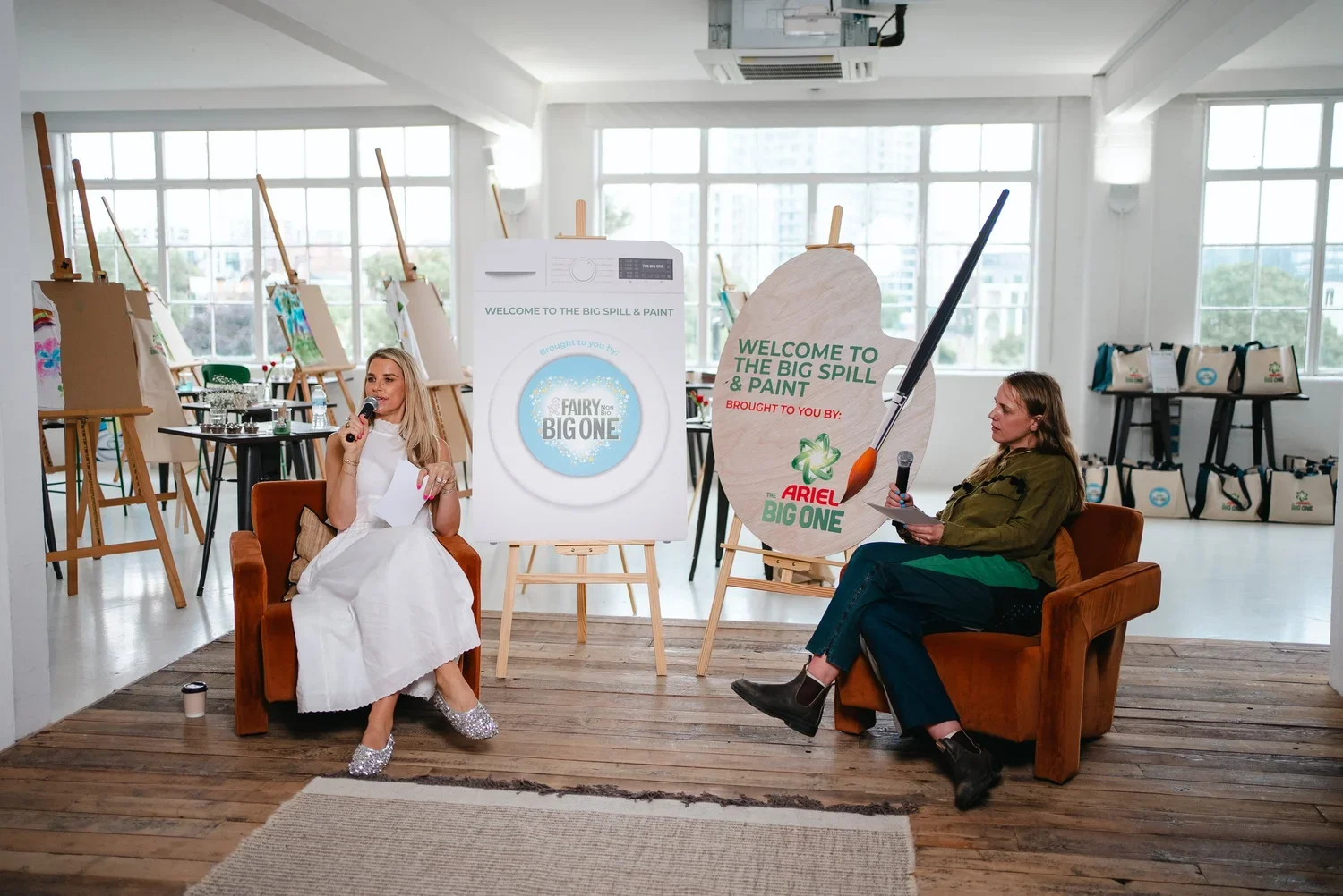 Two women seated in front of large posters and easels, engaging in a conversation or interview in an art studio with paintings on display and large windows in the background.