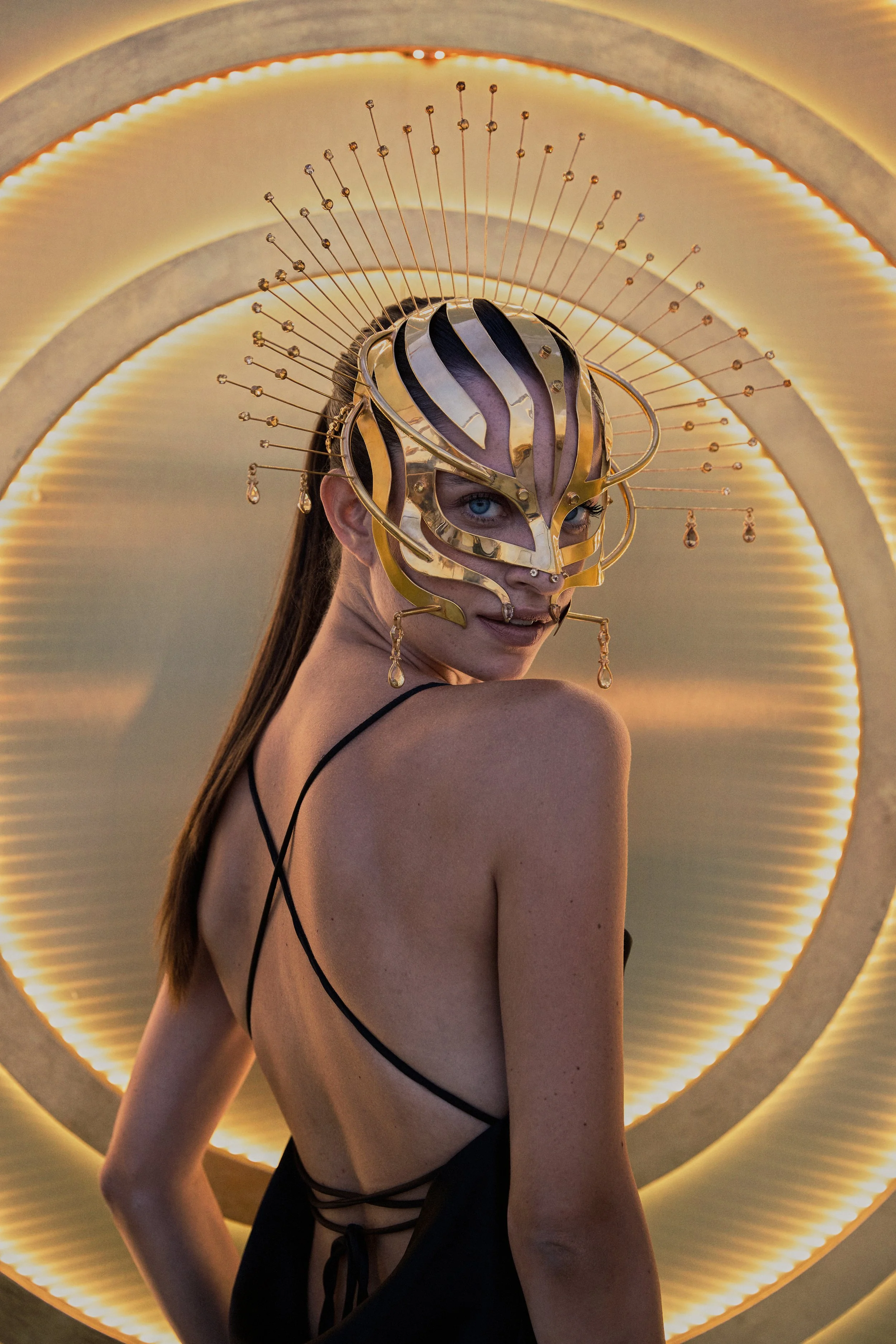 A woman with blue eyes and long brown hair is wearing an elaborate gold and purple mask with hanging jewelry. She is dressed in a black strappy outfit with an open back. The background features a series of illuminated concentric circles.