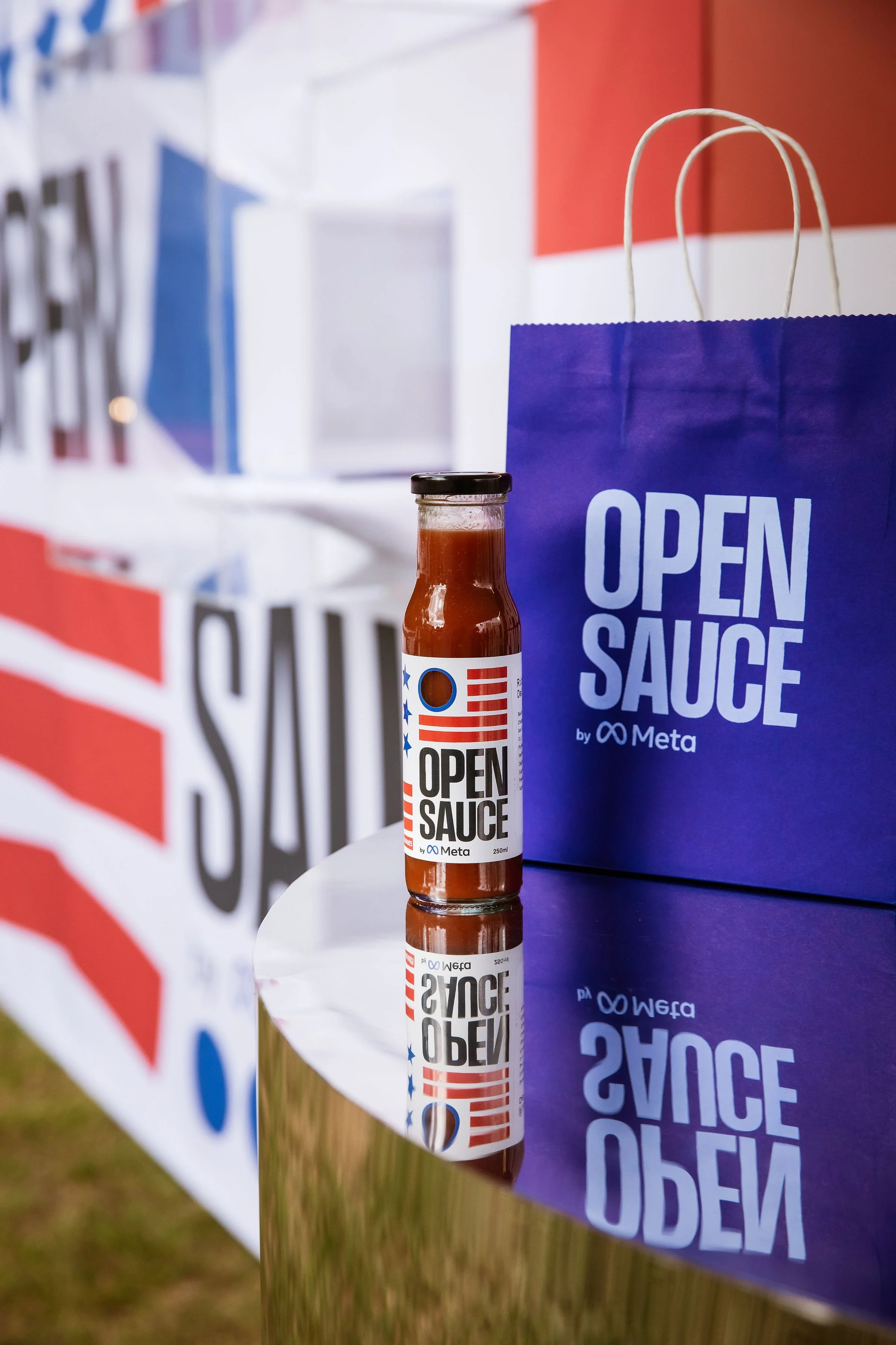 A bottle of sauce with a white label reading 'Open Sauce by Meta' next to a blue shopping bag with the same logo, placed on a reflective surface. In the background, there are promotional banners or signs.