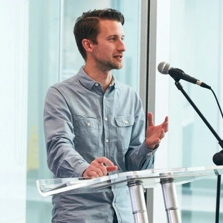 Young man in a blue button-up shirt speaking at a clear podium with a microphone in front of him.
