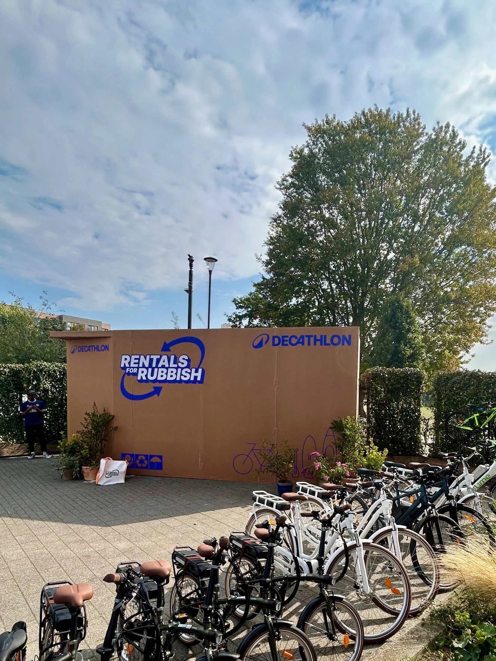 A row of bicycles parked outside on a cloudy day with a large tree, a building, and a sign that reads 'Rentals for Rubbish' in the background.