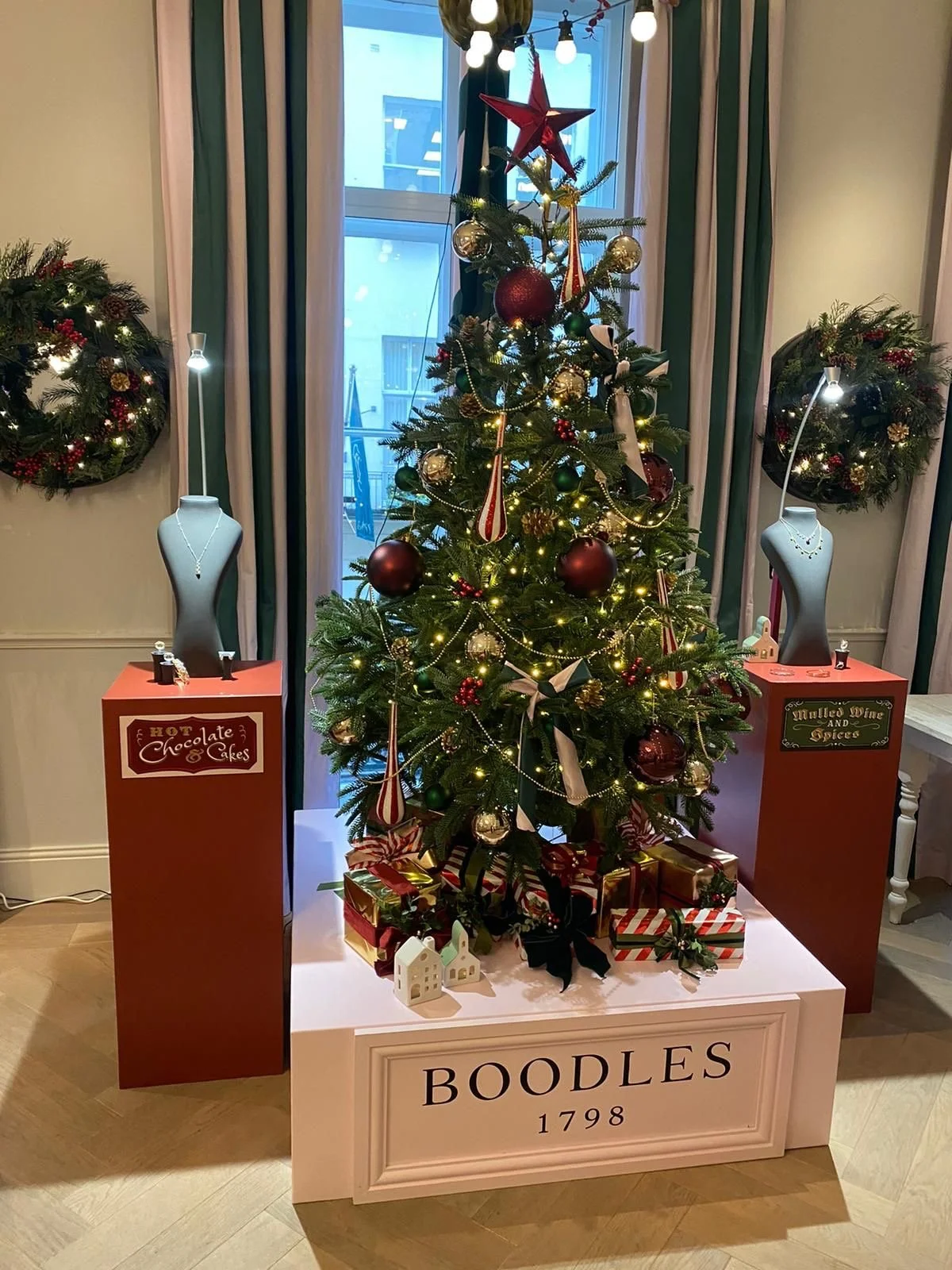Christmas tree decorated with gold and red ornaments, garland, ribbons, and lights, with wrapped gifts underneath, set on a white platform labeled "BOODLES 1798". Flanked by displays of jewelry on bust mannequins on orange pedestals, situated in a room with curtains, wreaths, and a window in the background.