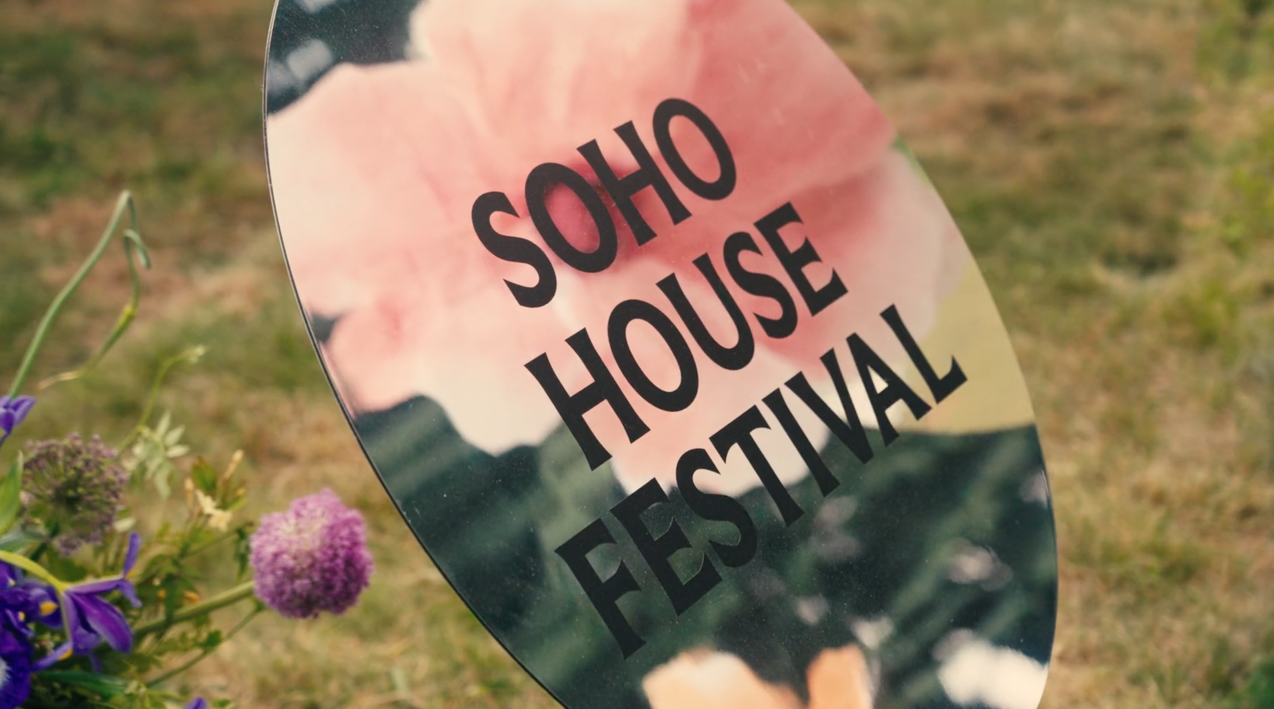 A reflective memorial mirror with the words 'SHOHOUSE FESTIVAL' printed on it, surrounded by flowers in a grassy outdoor setting.