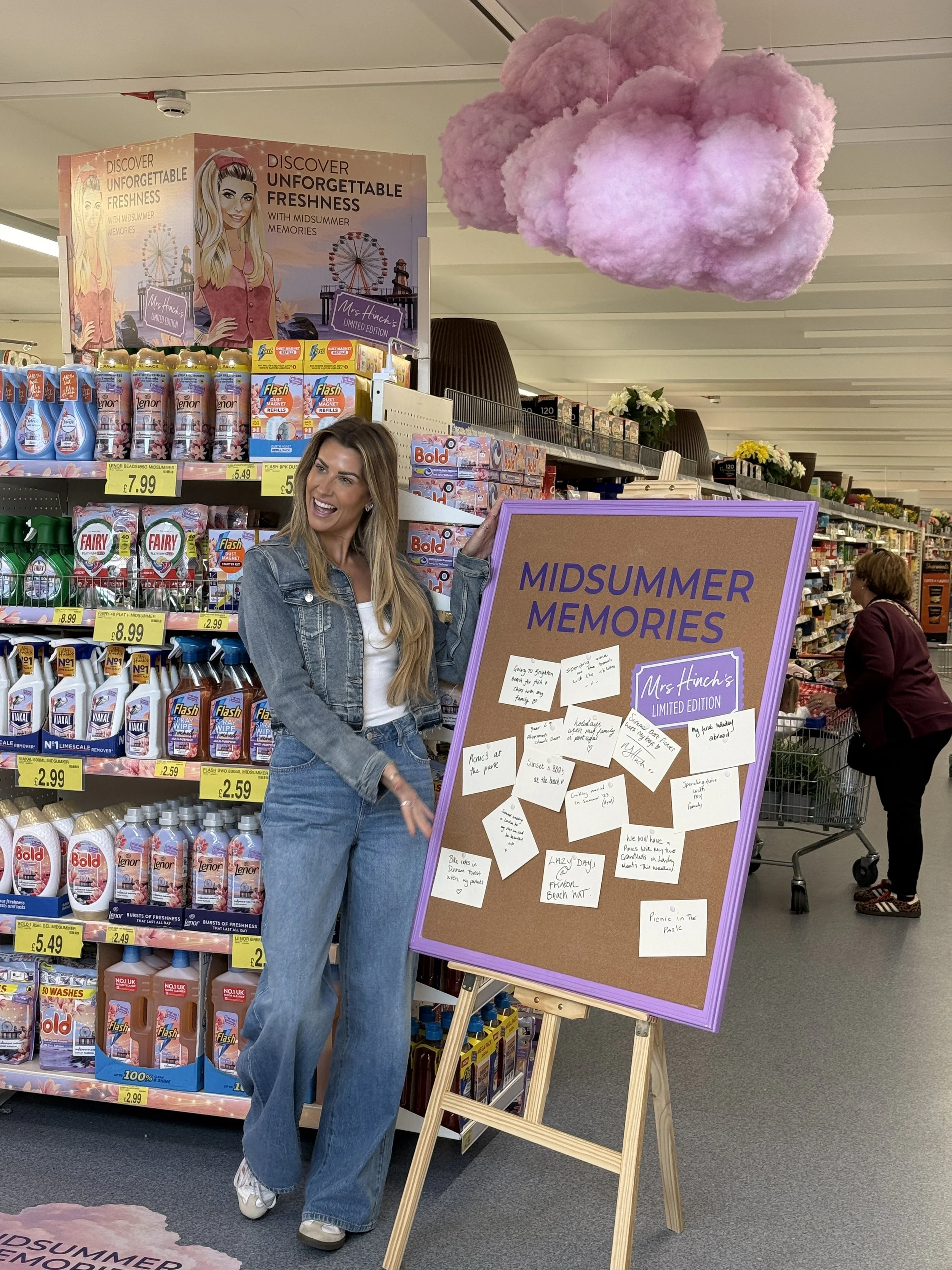 A woman standing in a store aisle next to a large purple board titled 'MIDSUMMER MEMORIES' with handwritten notes. She is smiling, wearing a denim jacket and jeans. There is a pink fluffy cloud decoration hanging from the ceiling and shelves stocked with cleaning supplies behind her.