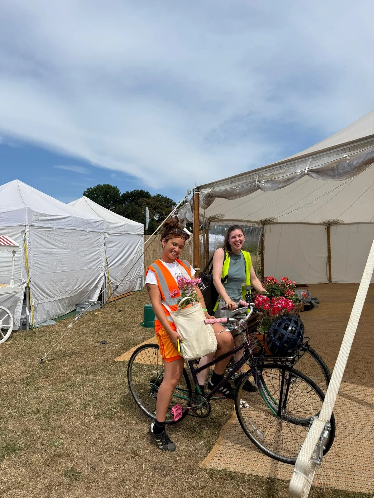 Two young women are standing outdoors with bicycles, laughing and smiling. They are wearing casual clothes and safety vests. The woman on the left has a white shirt with orange shorts, and the woman on the right has a grey shirt with black shorts. There are white tents and a large event tent in the background under a partly cloudy sky.