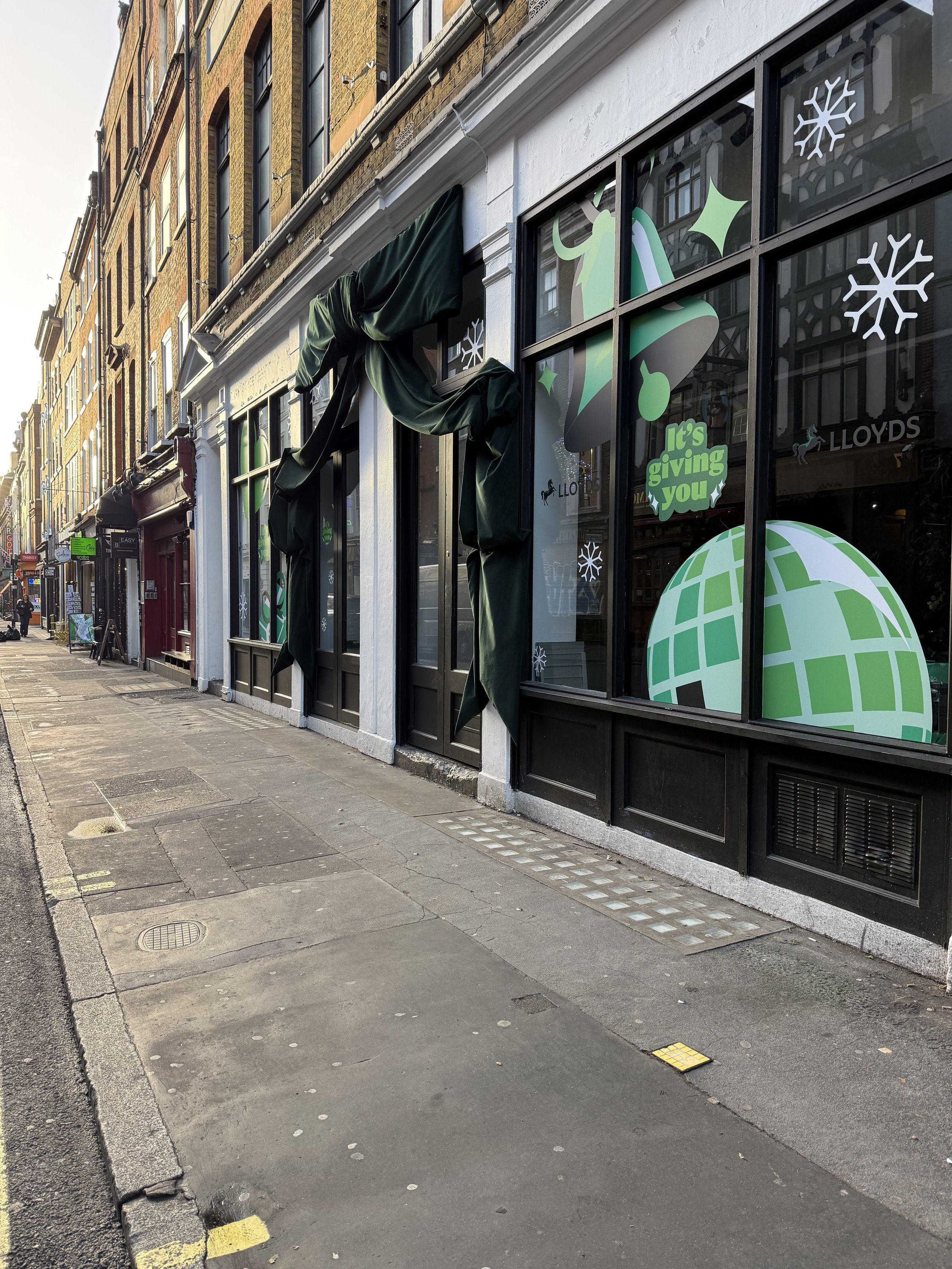 Empty city sidewalk in front of a storefront with Christmas decorations on the window and a green banner partially visible on the left side.