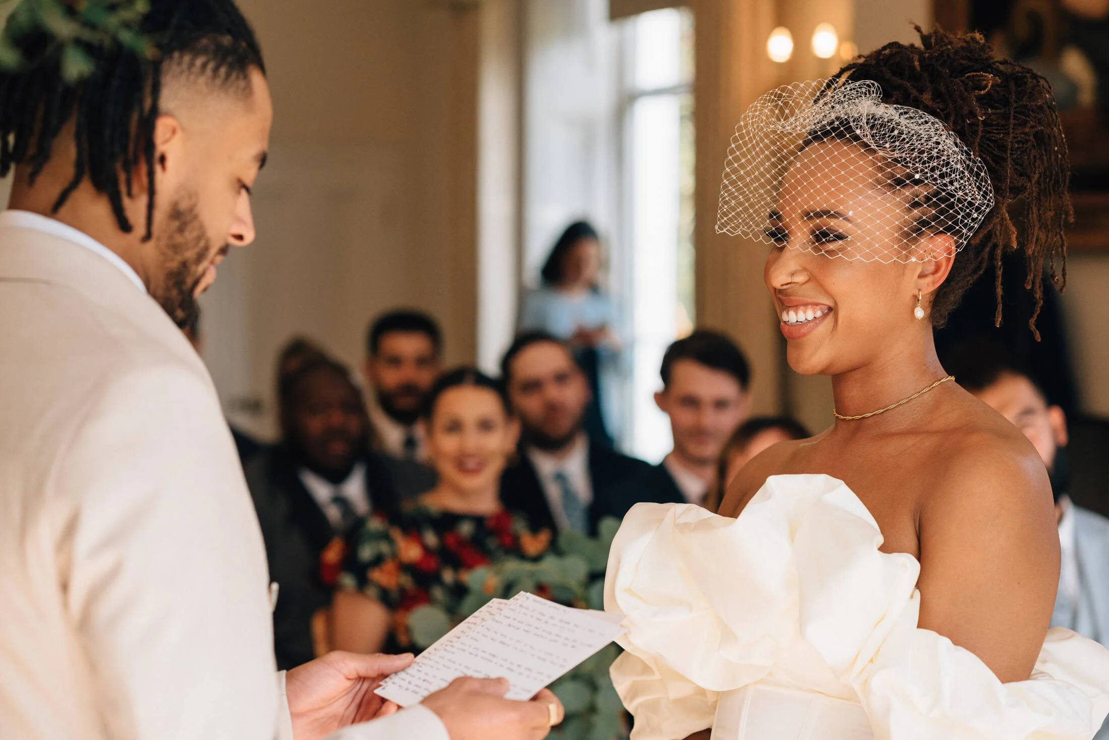 A woman receiving a marriage vows during a ceremony, wearing a white dress and a birdcage veil, smiling, with a crowd in the background.