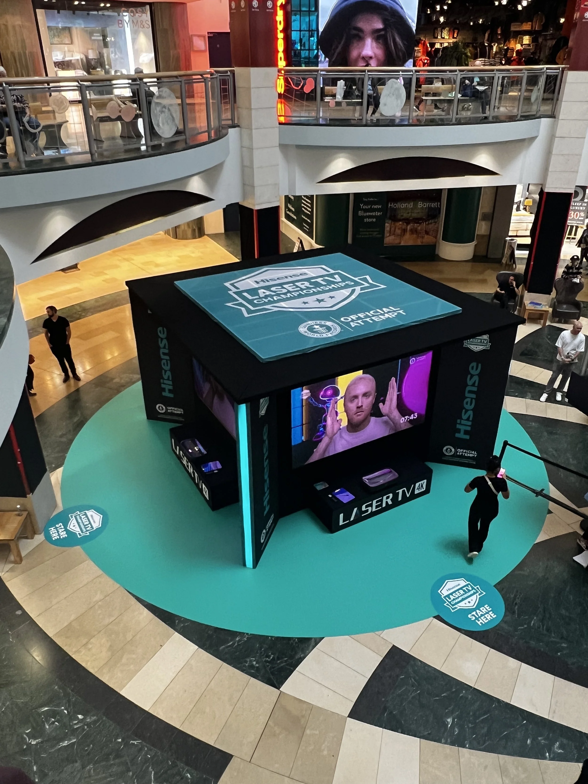 Indoor shopping mall with a central display promoting a NASCAR Las Vegas TV championship, featuring a large LED screen with a person on it, a smaller screen with a person's face, and a blue circular platform around the display with signs that say 'Share Here' and 'NASCAR TV'.