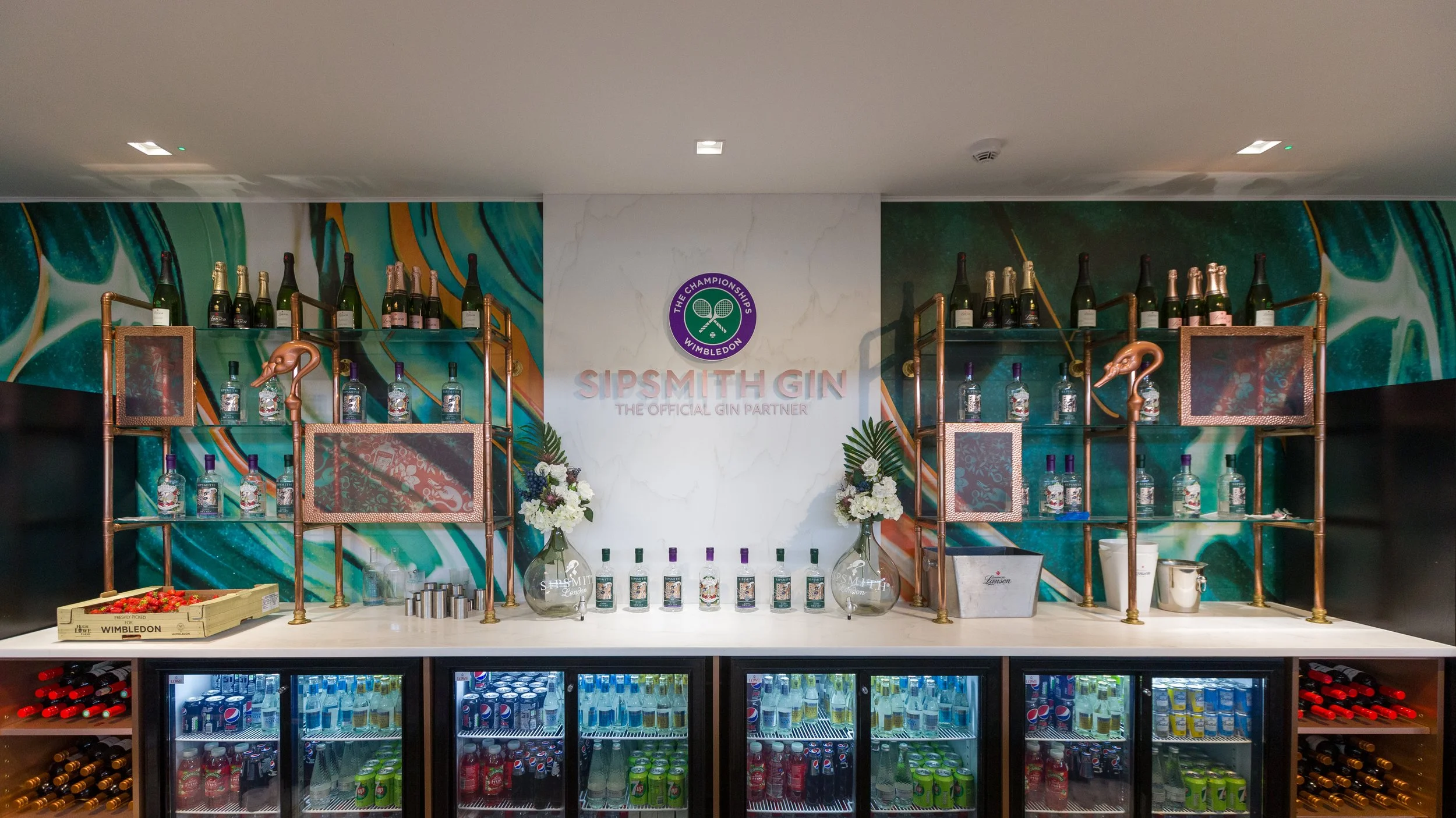 Display of bottles of Sipsmith Gin on copper shelving units with a colorful abstract mural background, a sign reading 'SIPSITH GIN' and 'THE OFFICIAL GIN PARTNER', and a refrigerator stocked with soft drinks below.