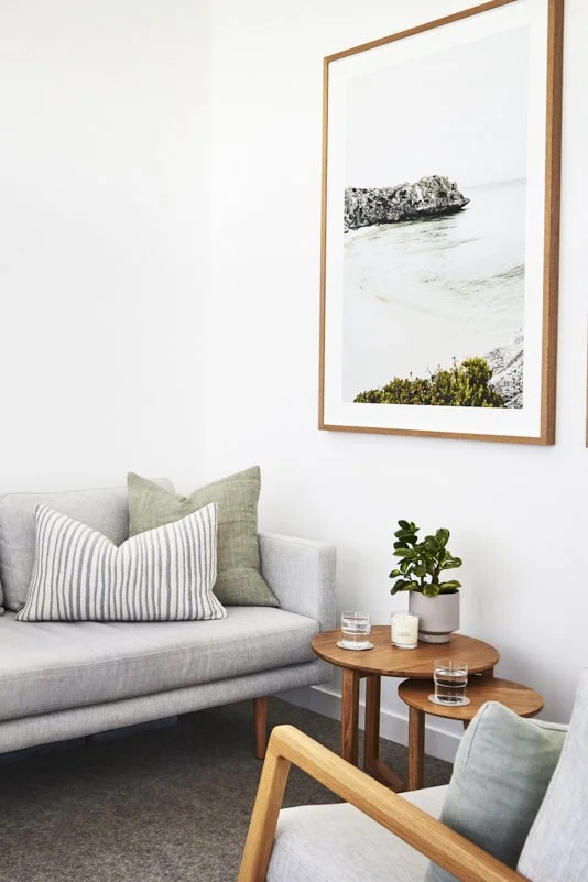 Living room with light gray sofa, green and striped pillows, wooden side tables with potted plant and glasses, light-colored armchair, and framed beach photograph on white wall.