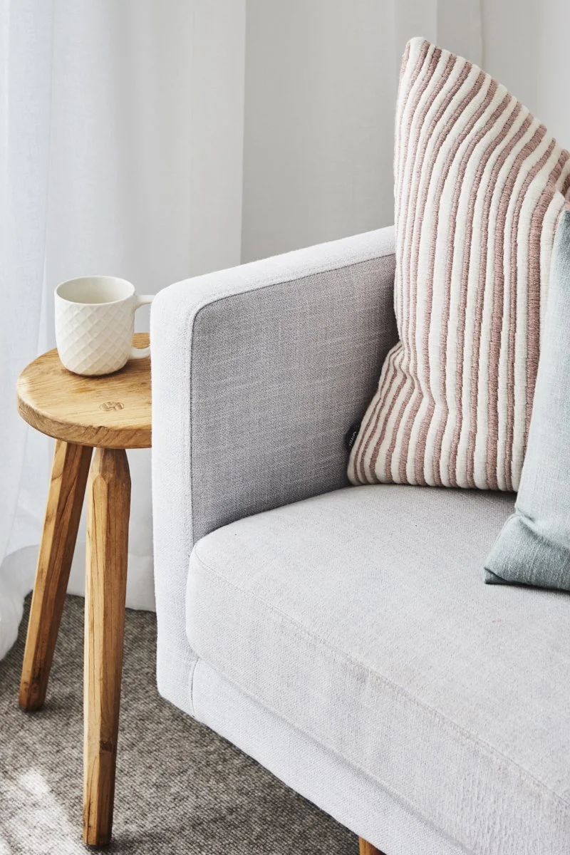 Close-up of a cozy living room corner featuring a light gray sofa with two decorative cushions, one striped pink and white, and the other solid gray. A small wooden side table with a natural finish holds a textured white mug. The background includes a white curtain and light-colored wall.