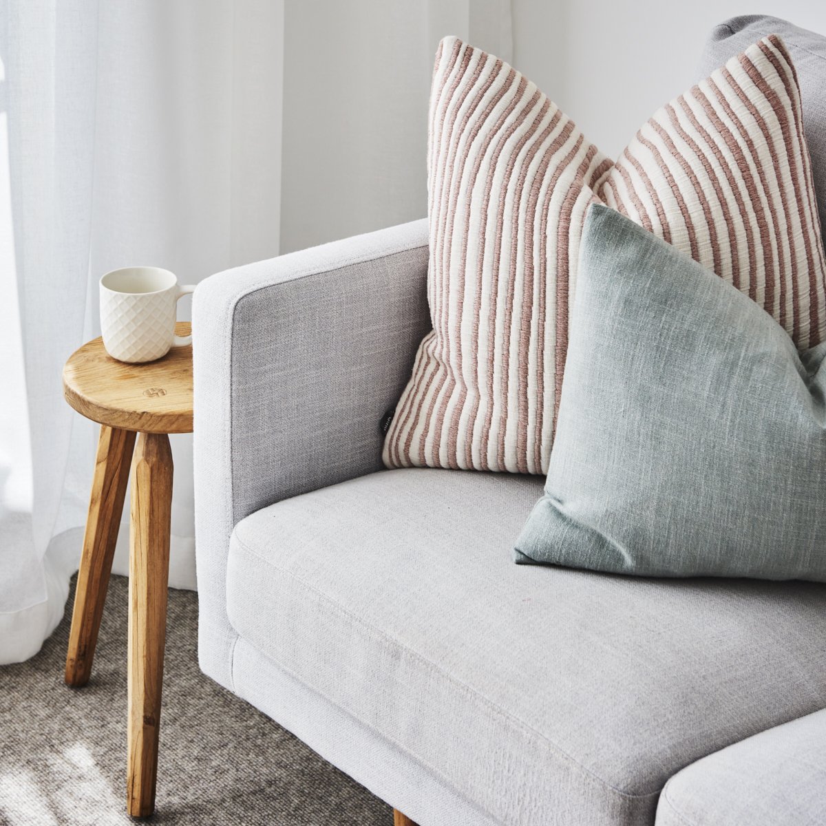 Close-up of a light gray sofa with a pink and white striped pillow, a light blue pillow, and a wooden side table with a white textured mug on it, next to white curtains.
