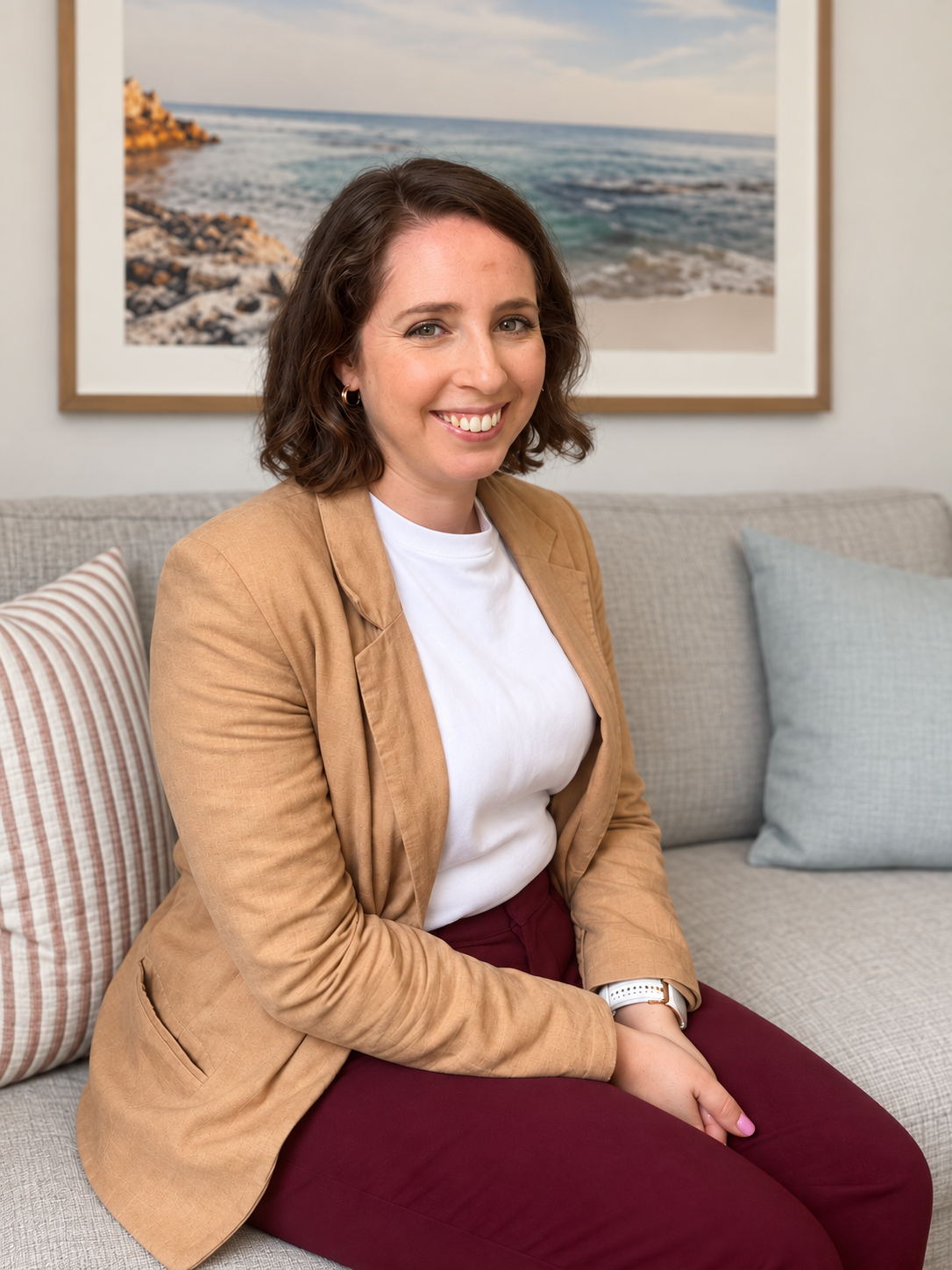 A woman with brown, curly hair, wearing a tan blazer, white t-shirt, and burgundy pants, sitting on a beige couch with patterned and light blue pillows, in a room with a framed beach scene photograph on the wall behind her.