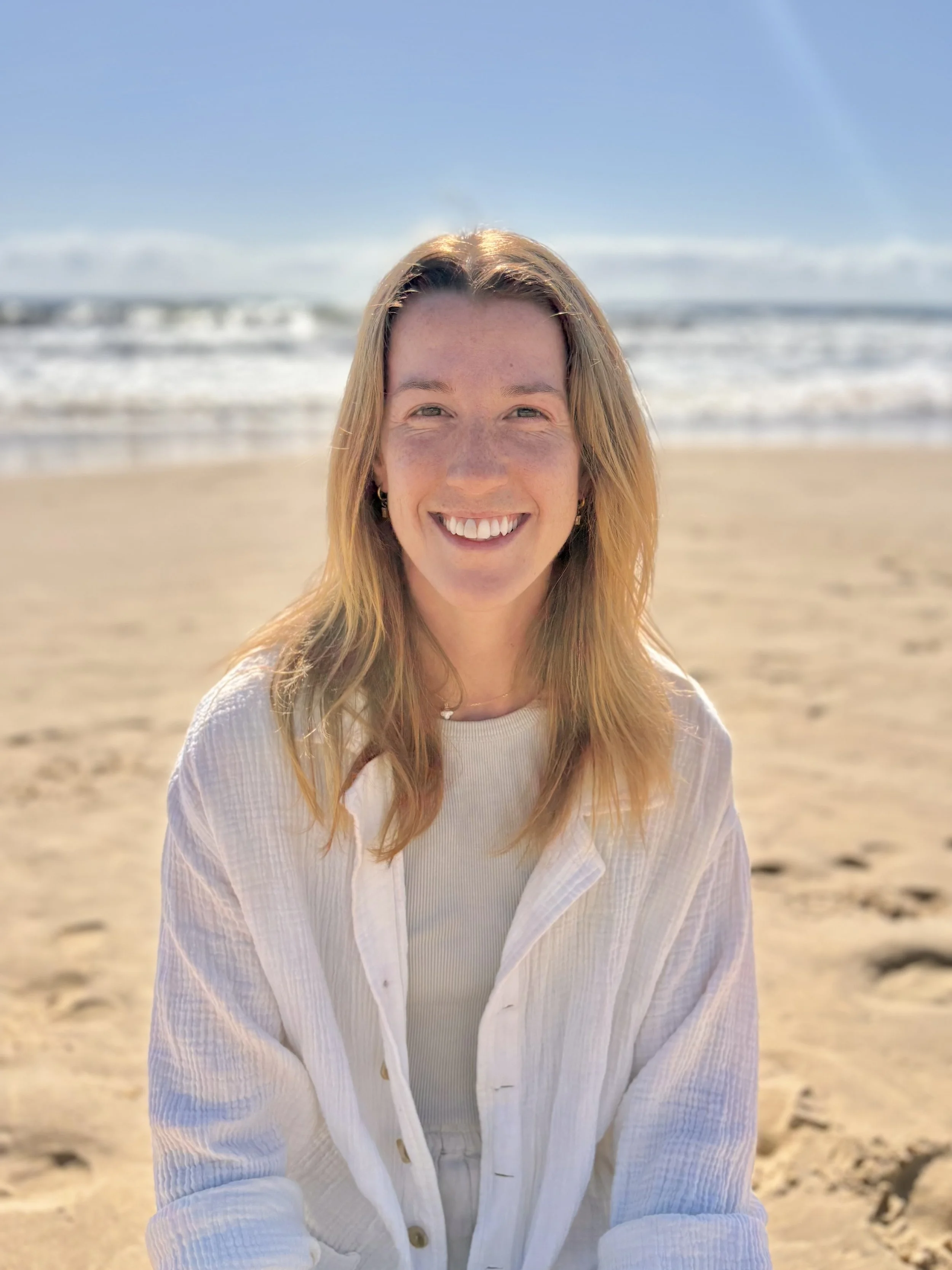 A smiling woman with long, light brown hair standing on a sandy beach with waves and blue sky in the background.