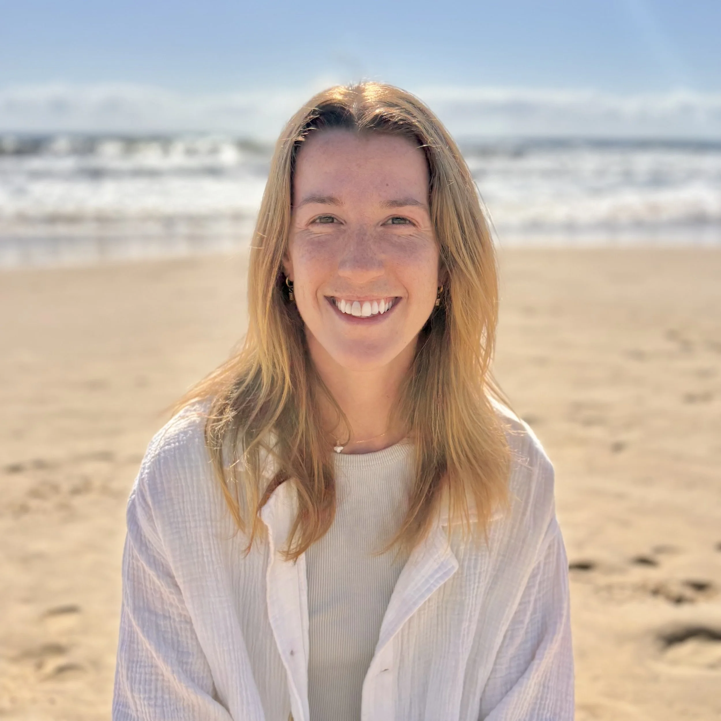 Close-up of a young woman with blonde hair smiling at the camera on a beach with waves in the background.