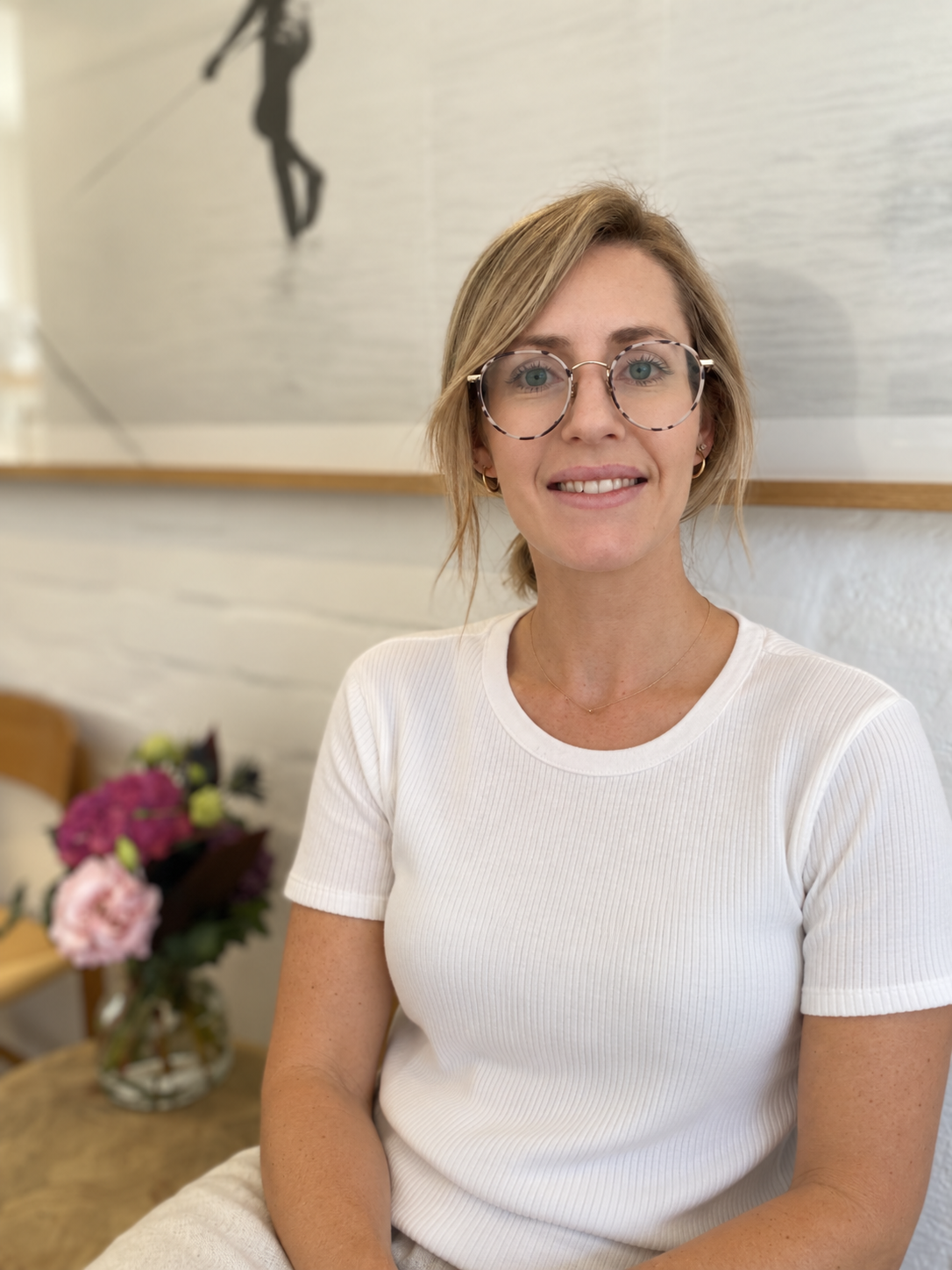 A woman with blonde hair and glasses smiling, sitting in a room with a flower arrangement on a table behind her, and a painting of a person climbing a mountain on the wall.