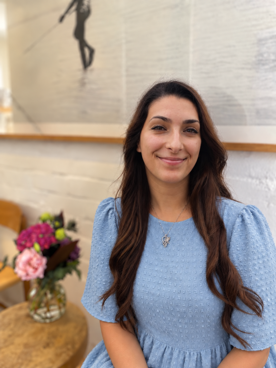 A woman with long brown hair smiling, wearing a light blue dress and a silver necklace, sitting in a room with a white brick wall and a floral arrangement on a table in the background.