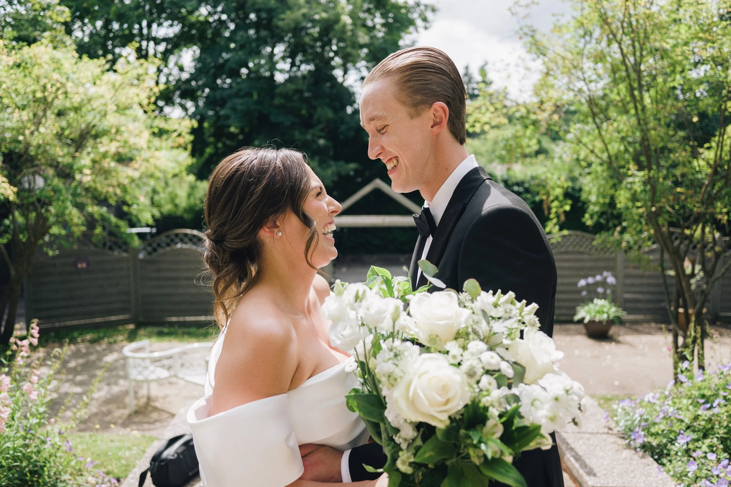 Bride holds a bouquet of flowers as she looks at her husband while the wedding photographer takes candid and natural photos of them