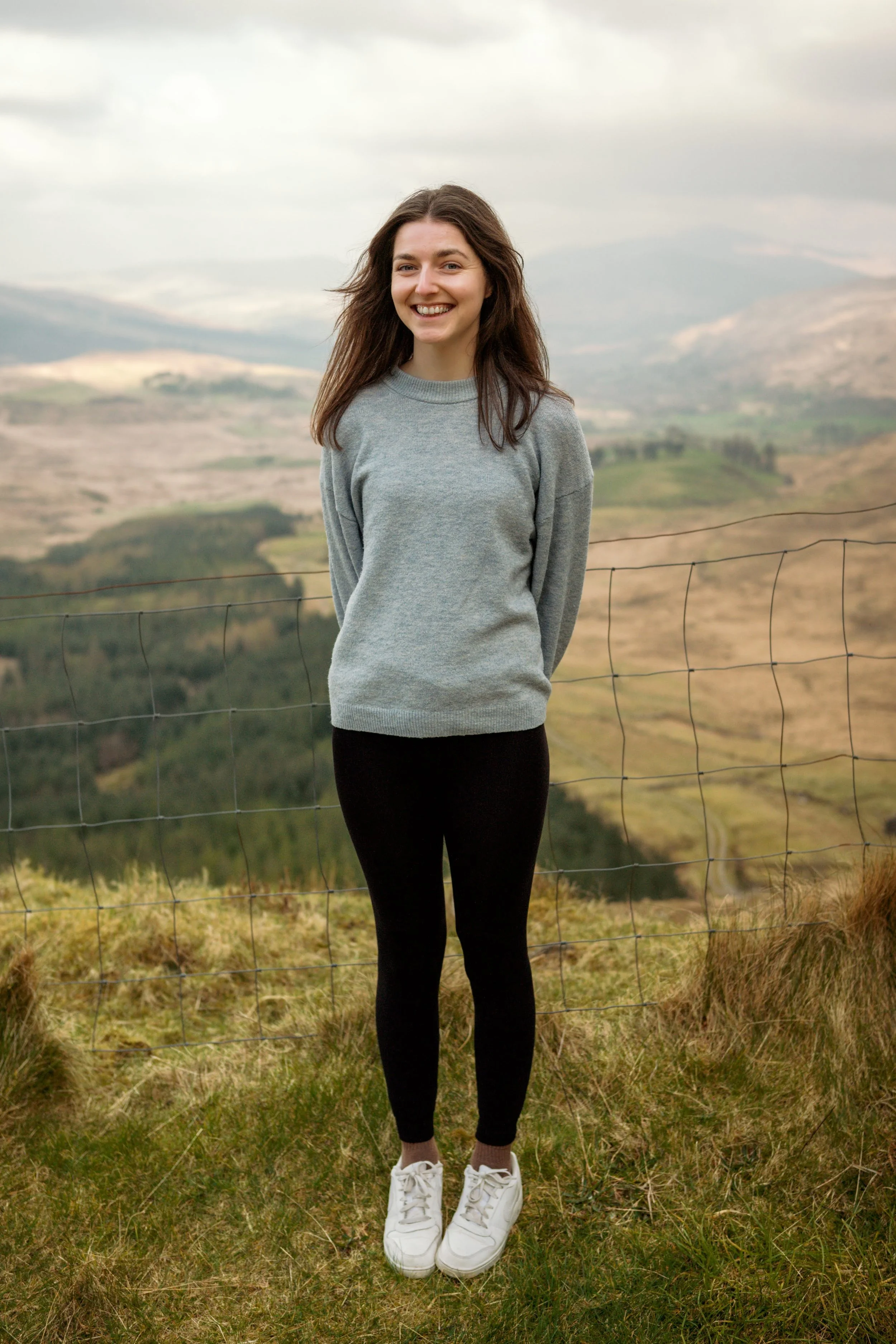 A young woman with long brown hair is smiling and standing outdoors in front of a wire fence, with rolling hills and a cloudy sky in the background.