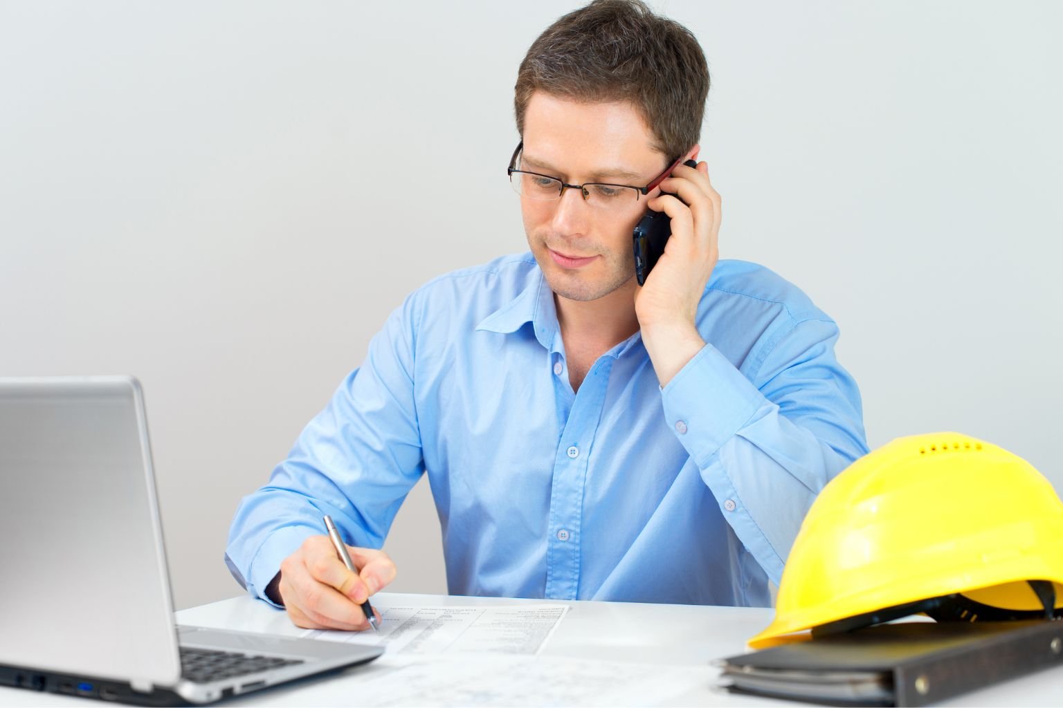 Electrician reviewing notes and discussing an electrical repair while working at a desk with a laptop and safety helmet.