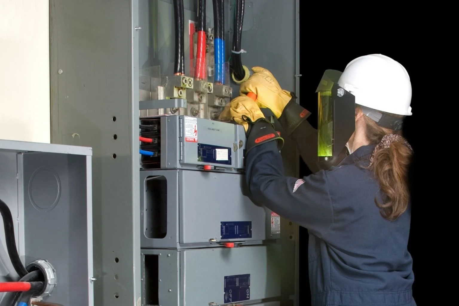 Electrician wearing protective gear inspecting electrical service equipment inside a commercial electrical panel.