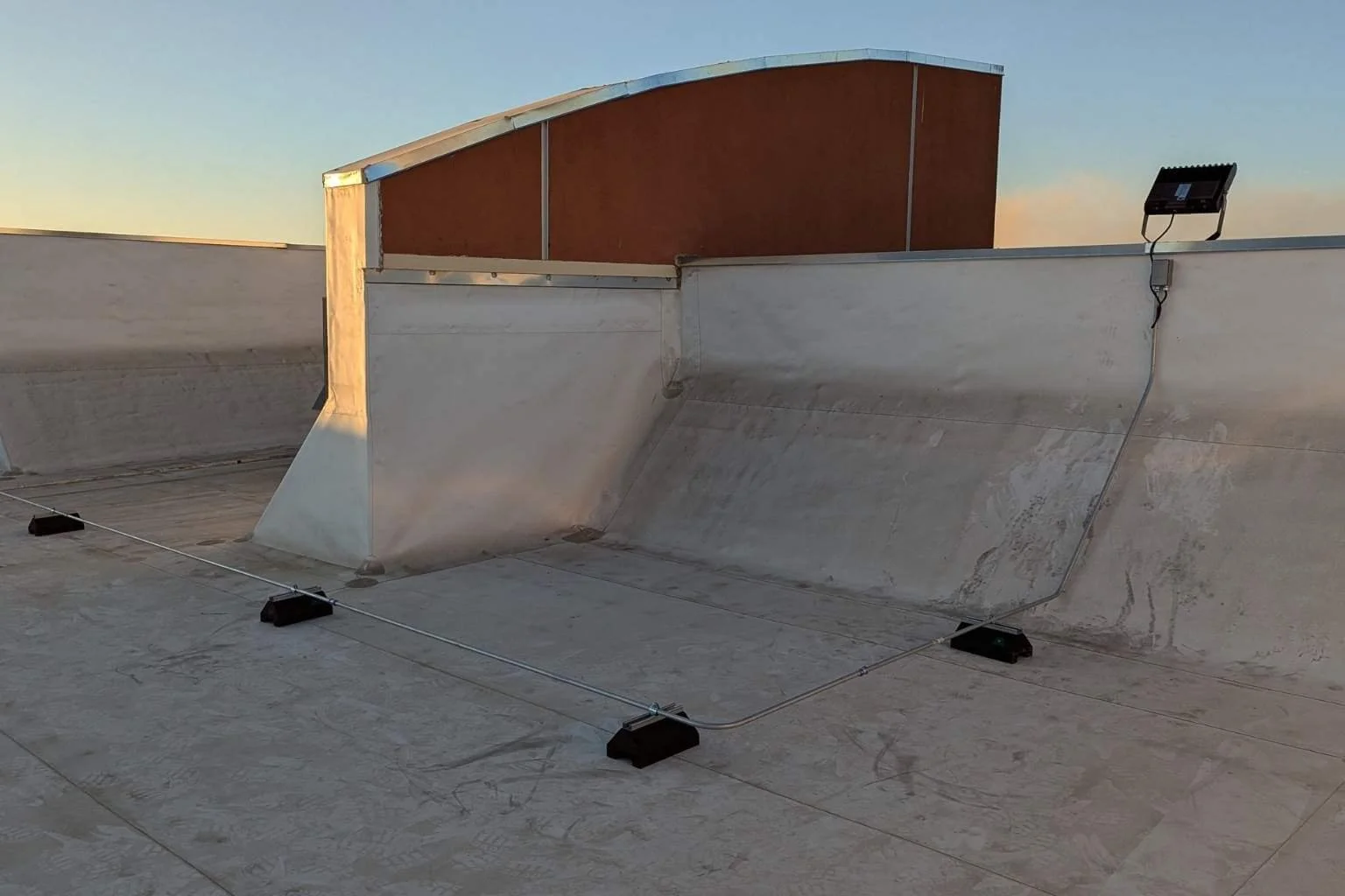 Electrical conduit and wiring installed on rooftop conduit supports to supply a new LED flood light at Cycle Gear in Victorville, California, providing power for exterior parking lot lighting.