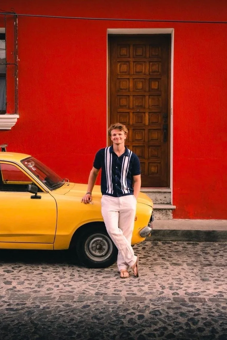 A young man standing beside a yellow car in front of a vibrant red building with a large wooden door and a small window with iron bars.