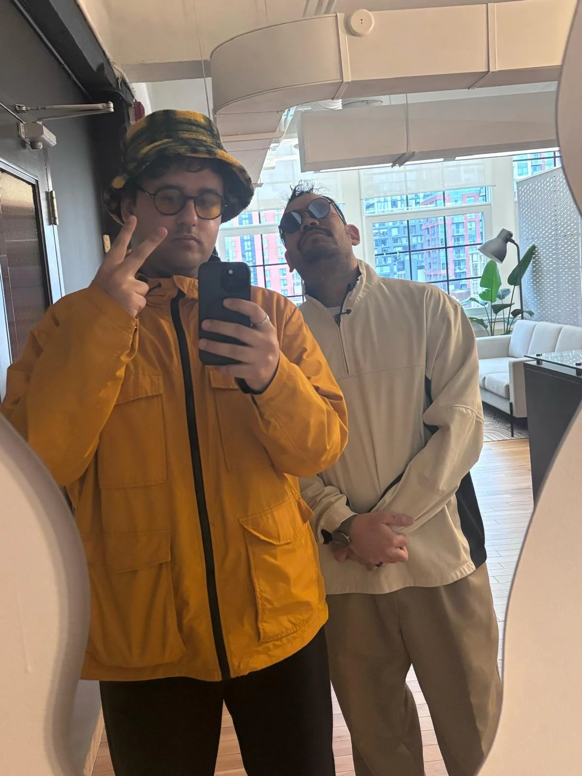 Two men taking a mirror selfie in a modern, brightly lit room with large windows and cityscape in the background. One man wears a yellow jacket, glasses, and a bucket hat, making a peace sign. The other wears a beige jacket and sunglasses.