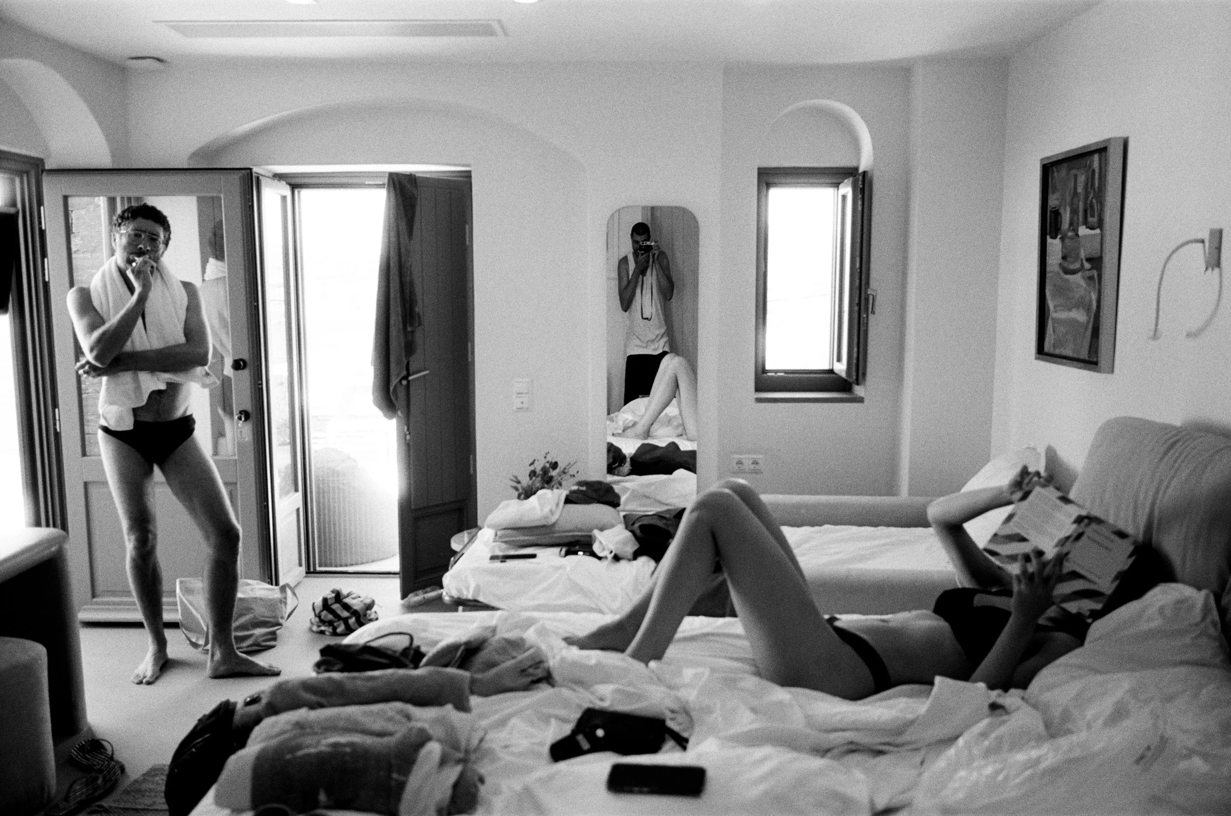 A black and white photo of a cluttered bedroom with two beds, a person lying on one bed reading a magazine, and two women standing and taking photos; one near the door and the other in the mirror, with the room open to outside.