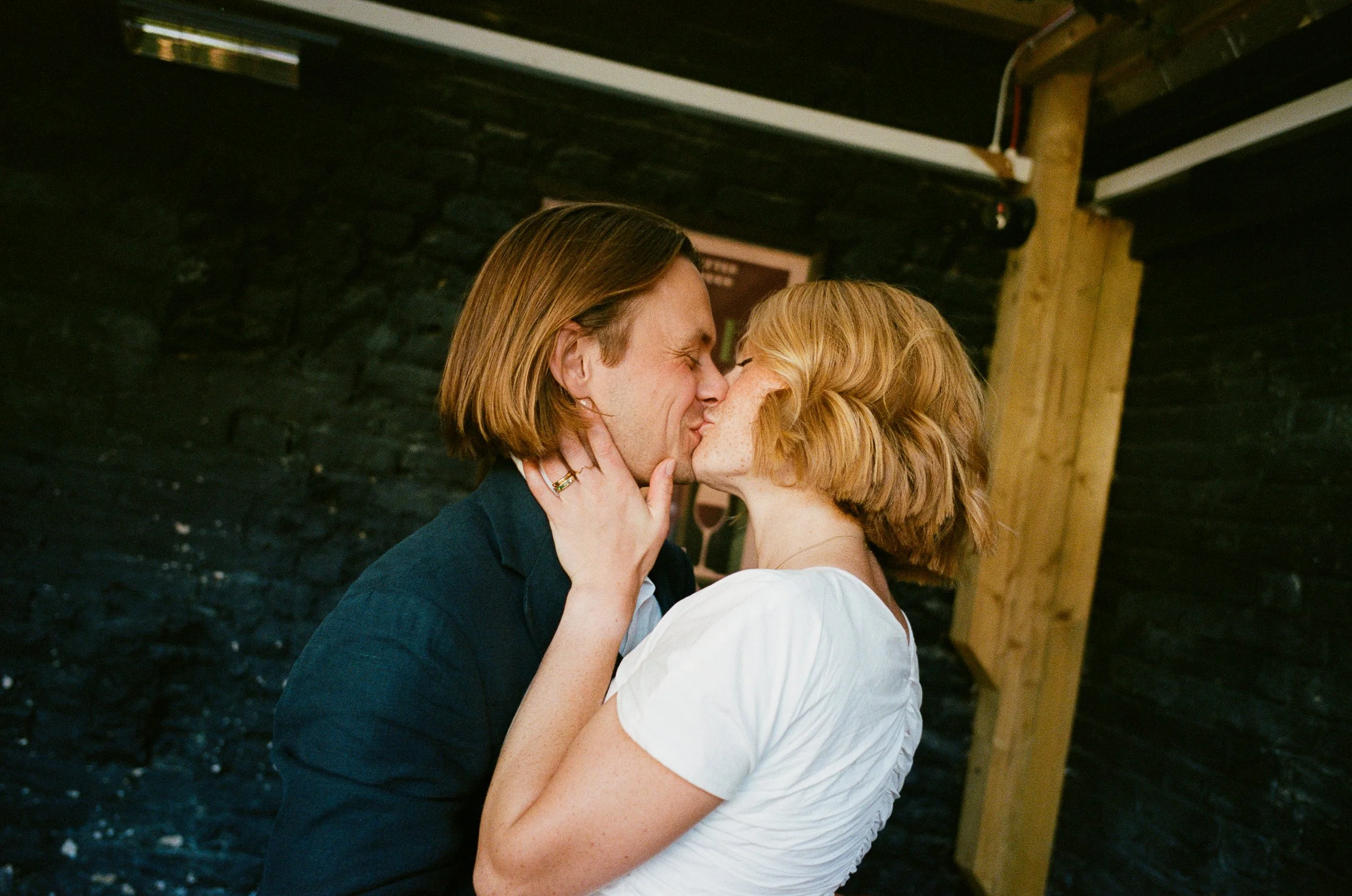 A couple sharing a kiss indoors, with a black brick wall and wooden beams in the background.