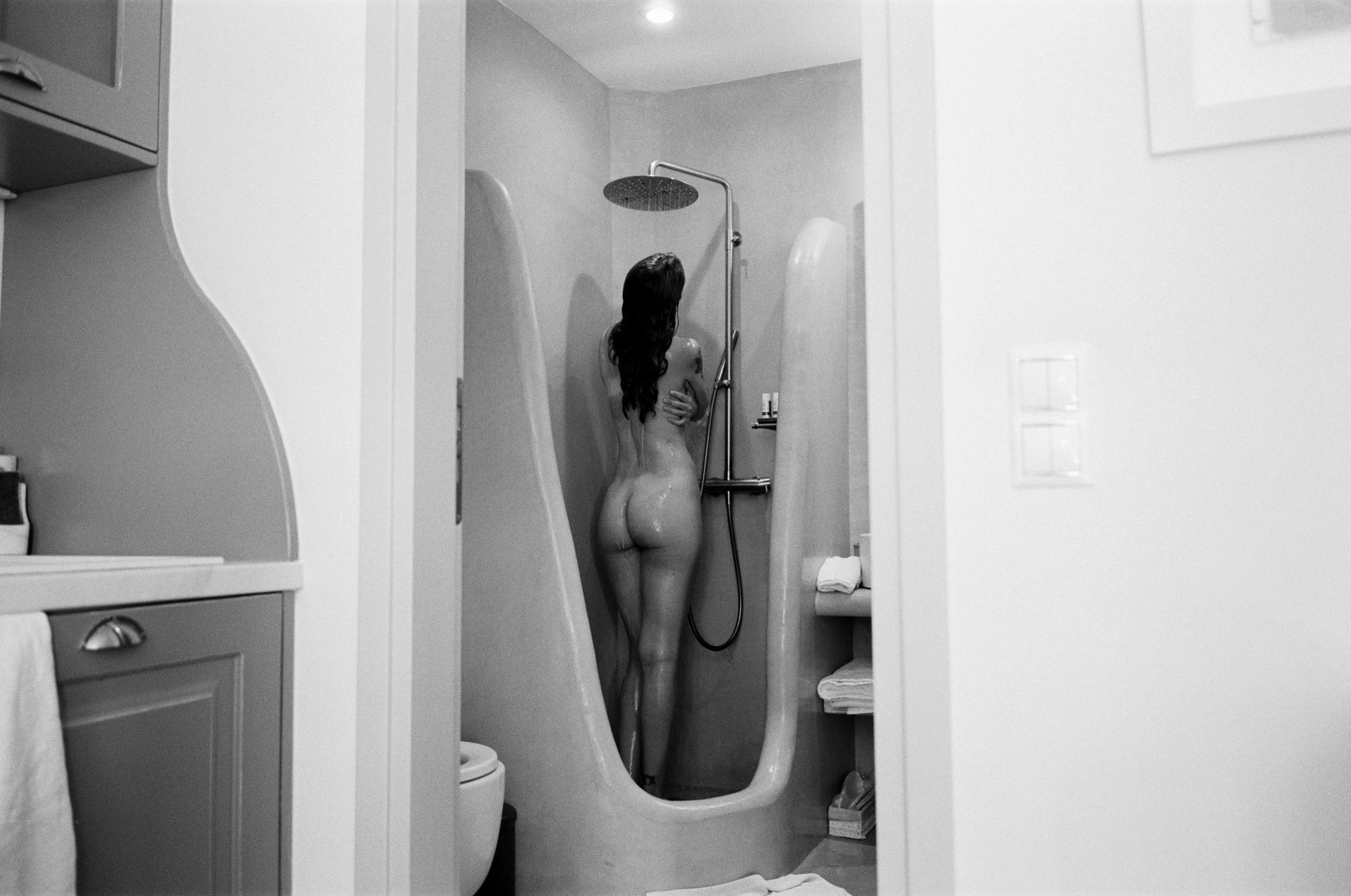 A woman with long hair taking a shower in a small bathroom with a shower head mounted on the wall. The scene is in black and white.