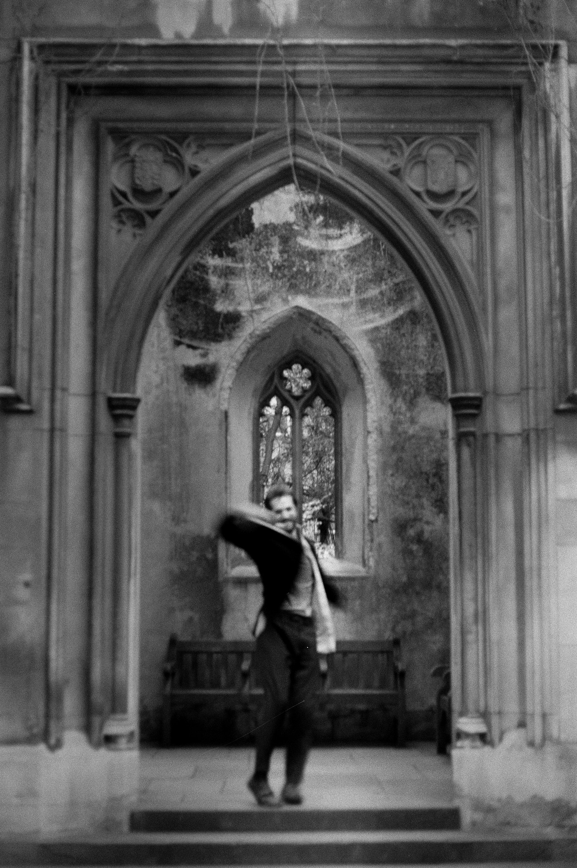Black and white photo of a man in a suit and hat walking down a corridor in front of a Gothic-style arched window, with stone walls and elaborate architectural details.
