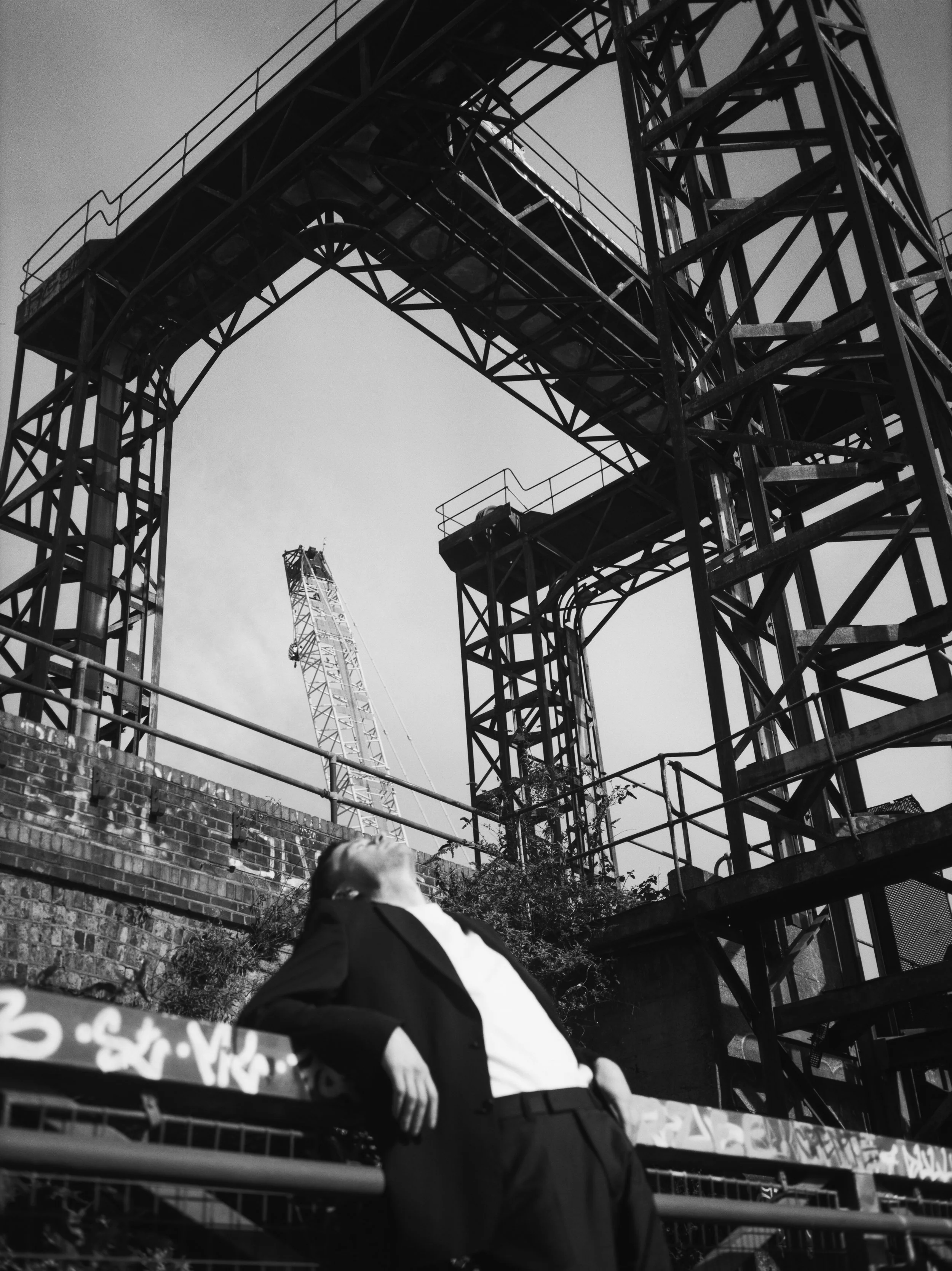 A man in a suit seated near a construction site with scaffolding and cranes in the background, viewed from below.