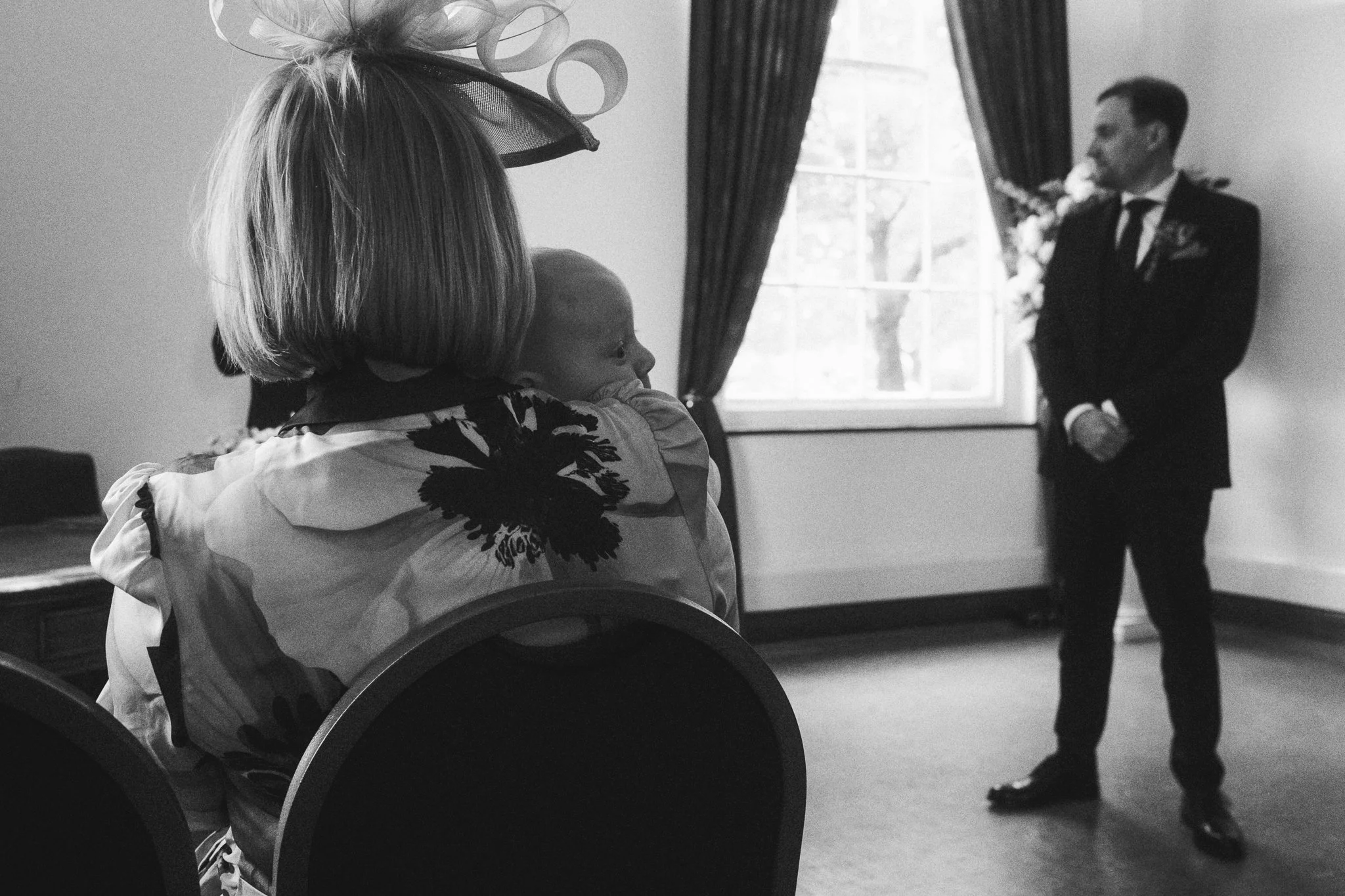 A woman with large statement earrings and a large fascinator hat in a floral pattern looks at a man in a suit and tie standing near a window. The photo is black and white.
